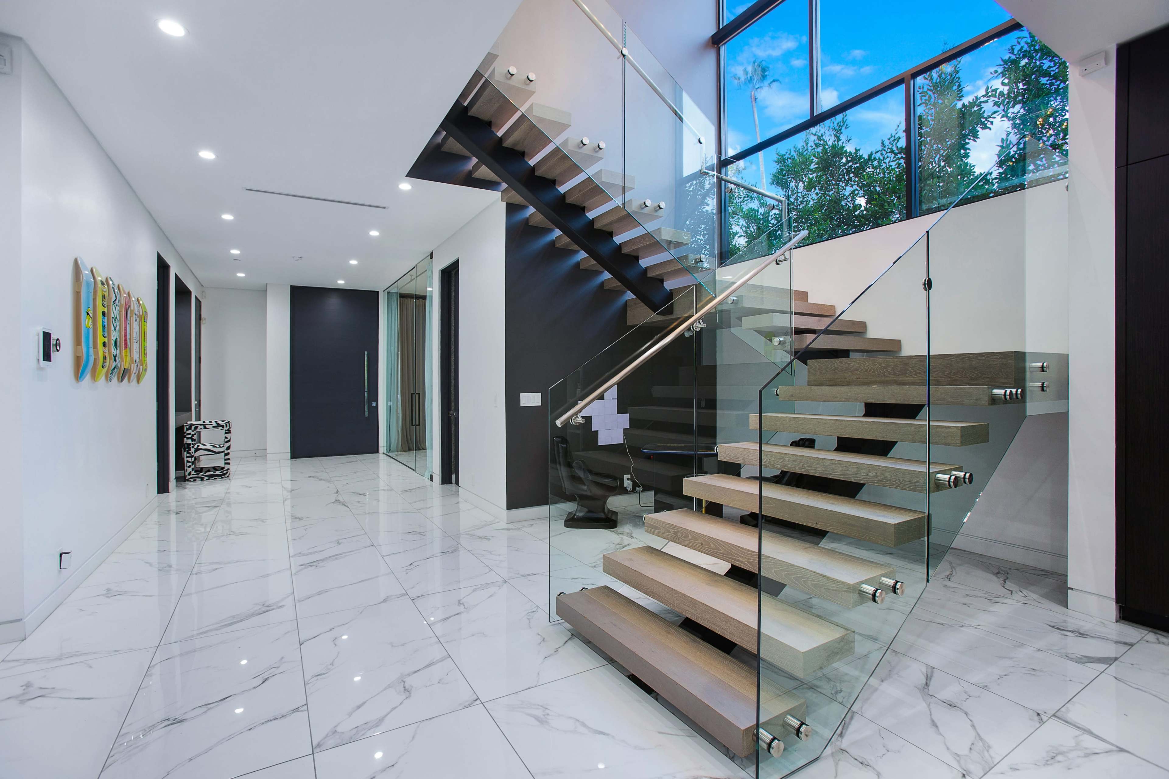 The image shows a modern entryway with a glass-walled staircase, marble flooring, and large windows allowing natural light.