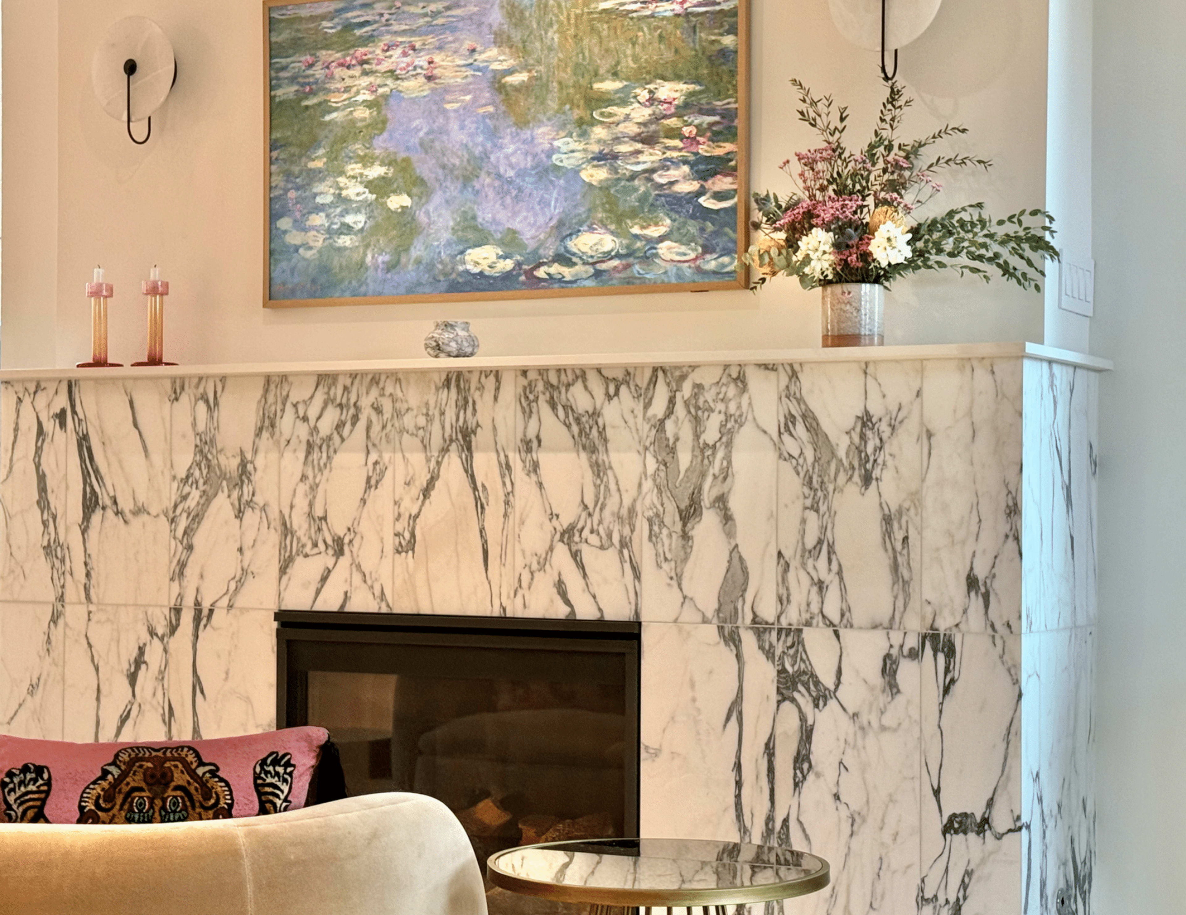 The image features a living room corner with a marble fireplace, a floral painting above it, and decorative items, including candles and a vase of flowers.