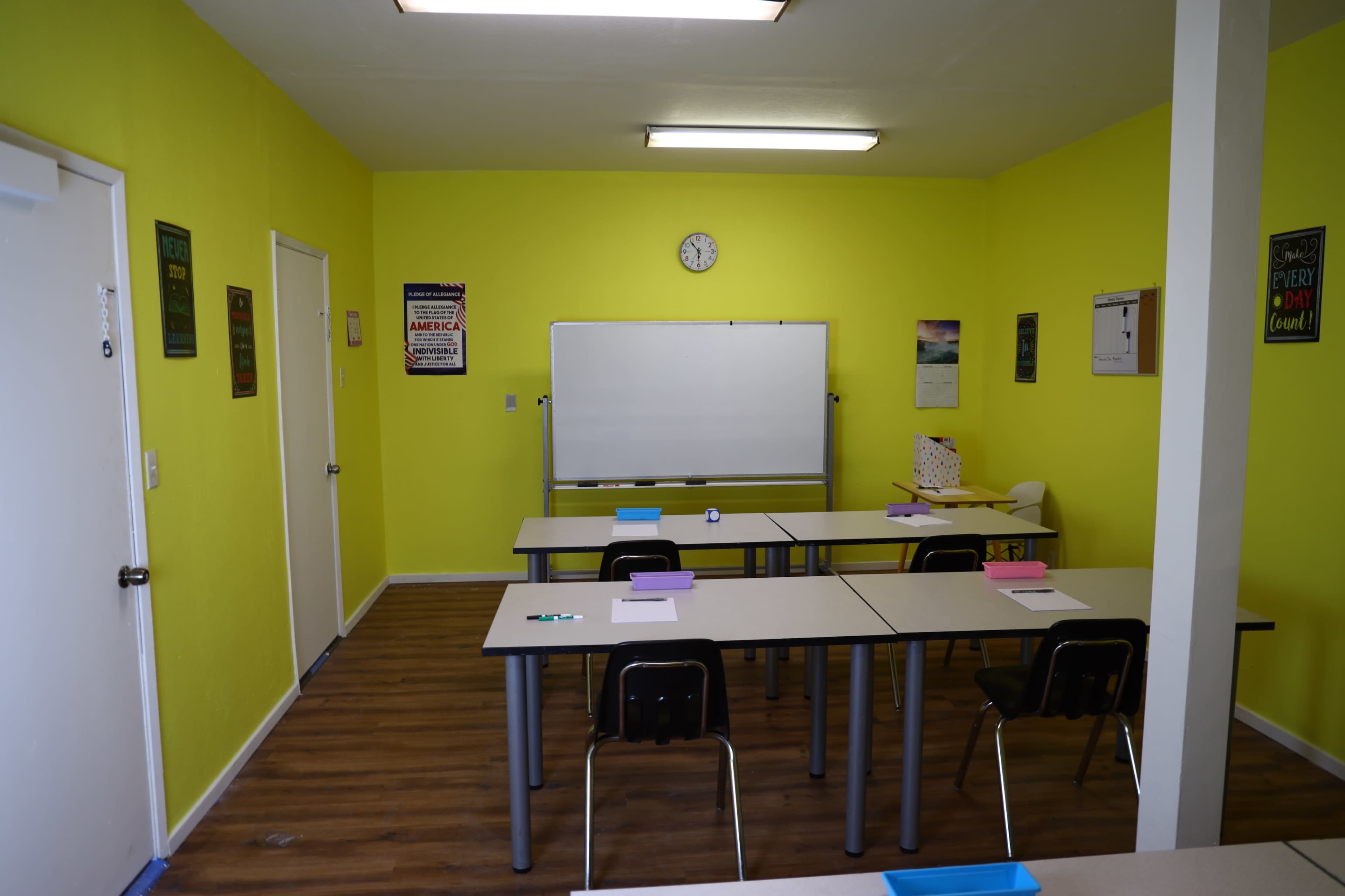 The image shows a brightly colored classroom with green walls, a whiteboard at the front, and several tables arranged with chairs.