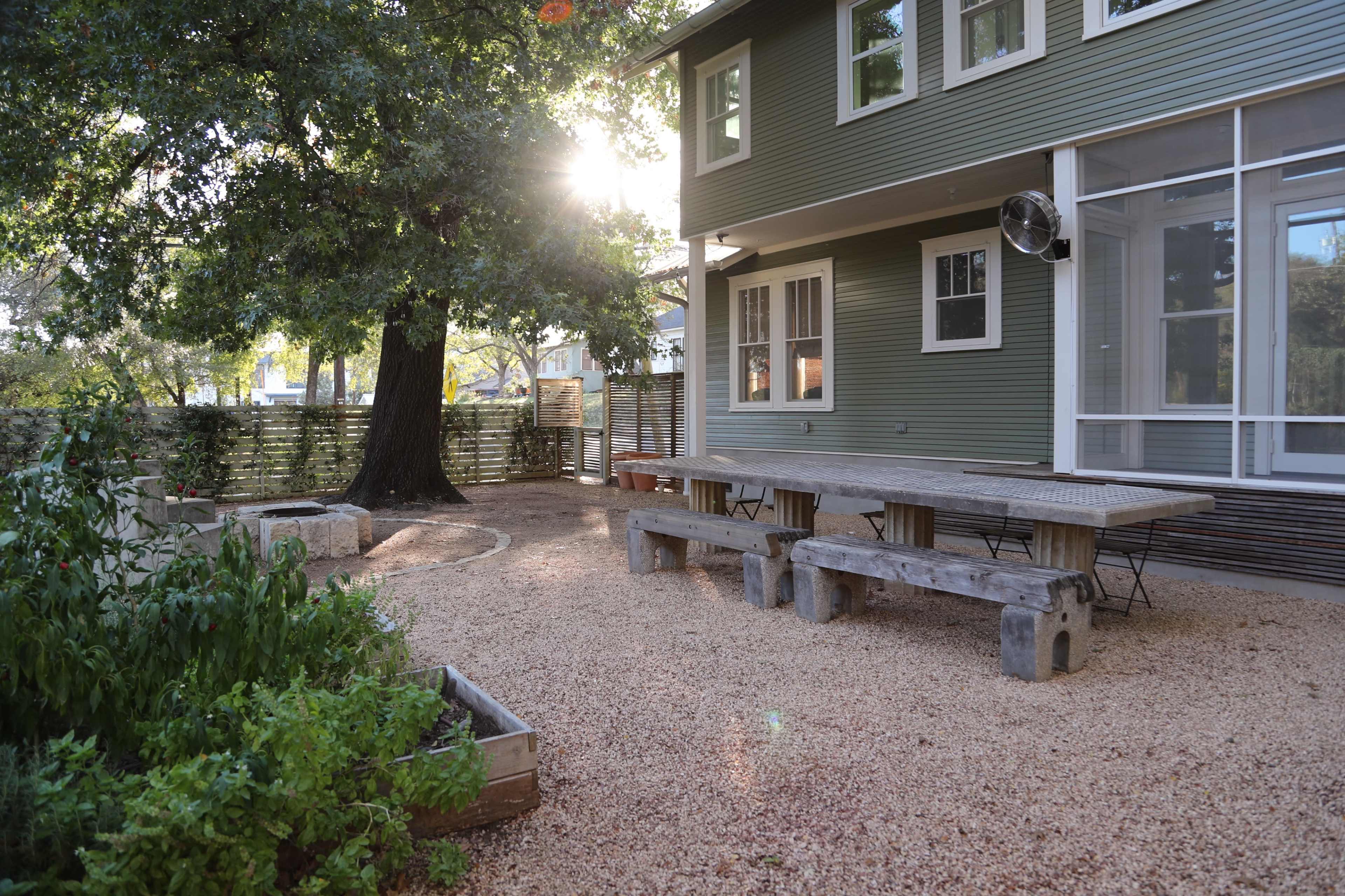 The image shows a backyard with a large wooden table surrounded by gravel, a garden bed with plants, and a two-story house with large windows.