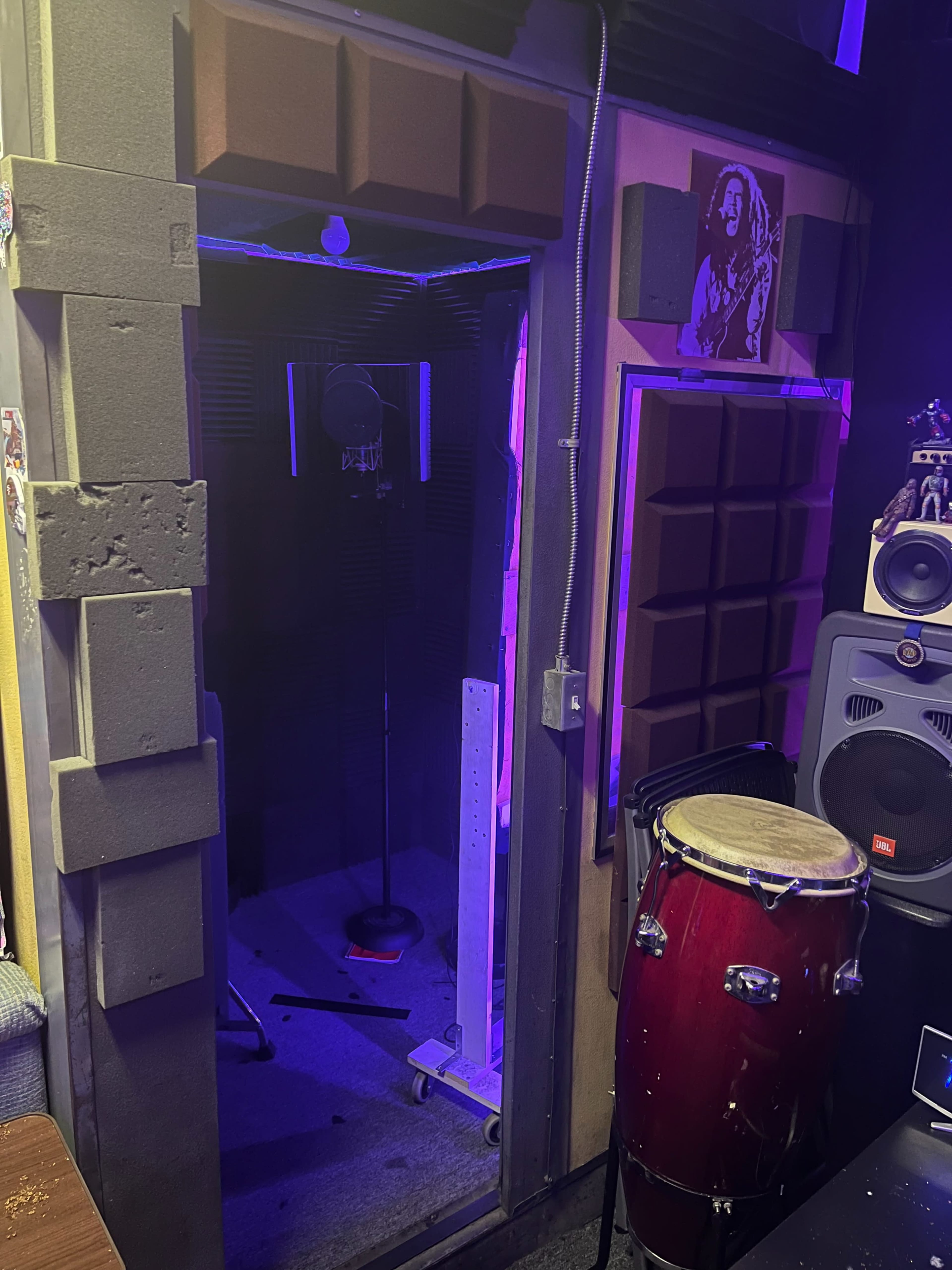A small recording studio booth with a microphone on a stand, surrounded by soundproof panels and illuminated by purple lights, alongside a conga drum and audio equipment.