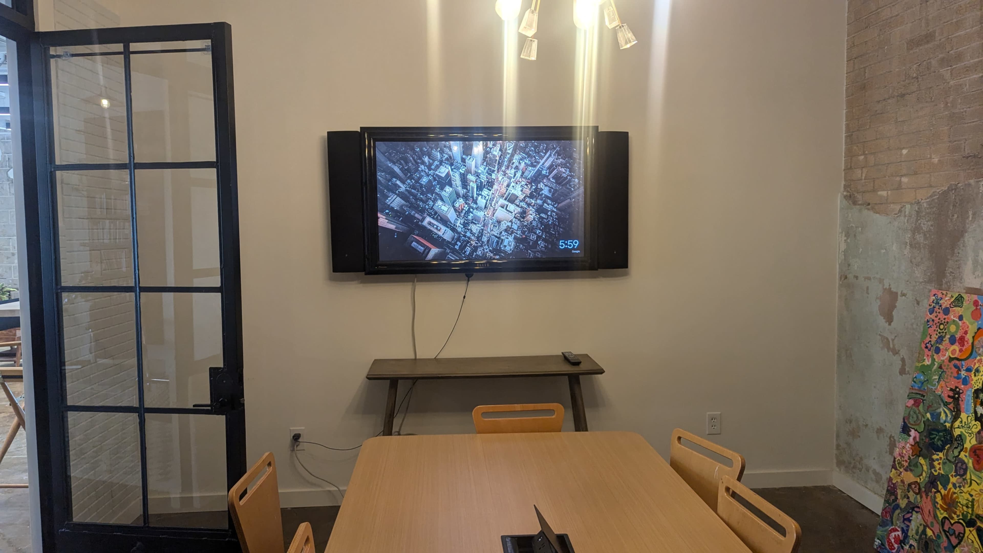 A modern meeting room features a wooden table surrounded by chairs, with a large wall-mounted television displaying a cityscape, and a glass door leading outside.