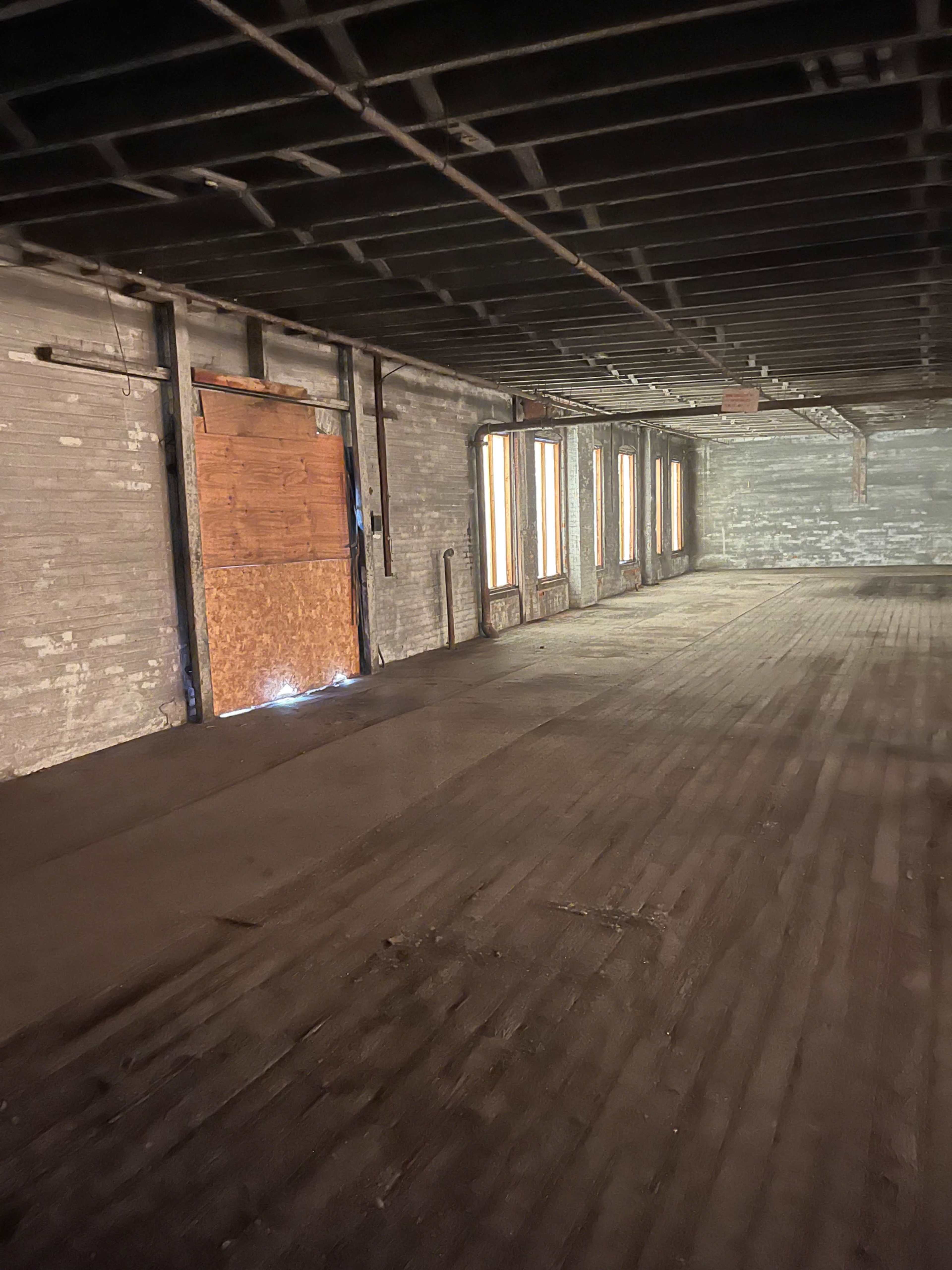 The image shows an empty, dimly lit room with wooden flooring and a boarded-up doorway, framed by several windows on one side.