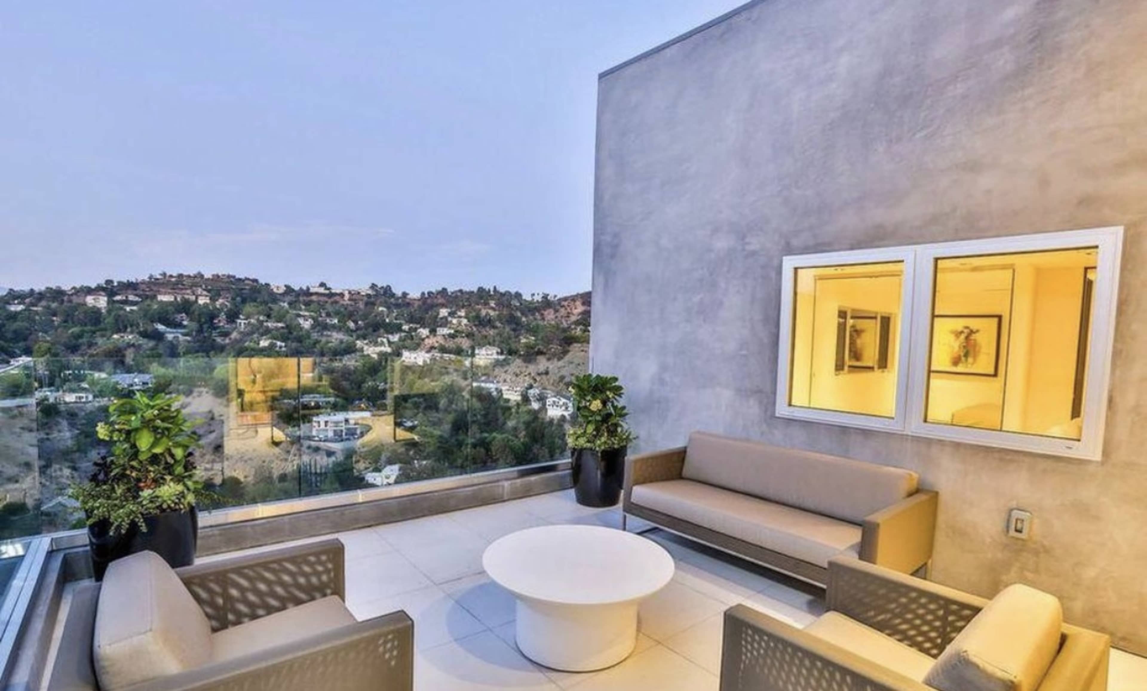 A modern patio features a round table surrounded by a sectional sofa and potted plants, overlooking a hilly landscape.