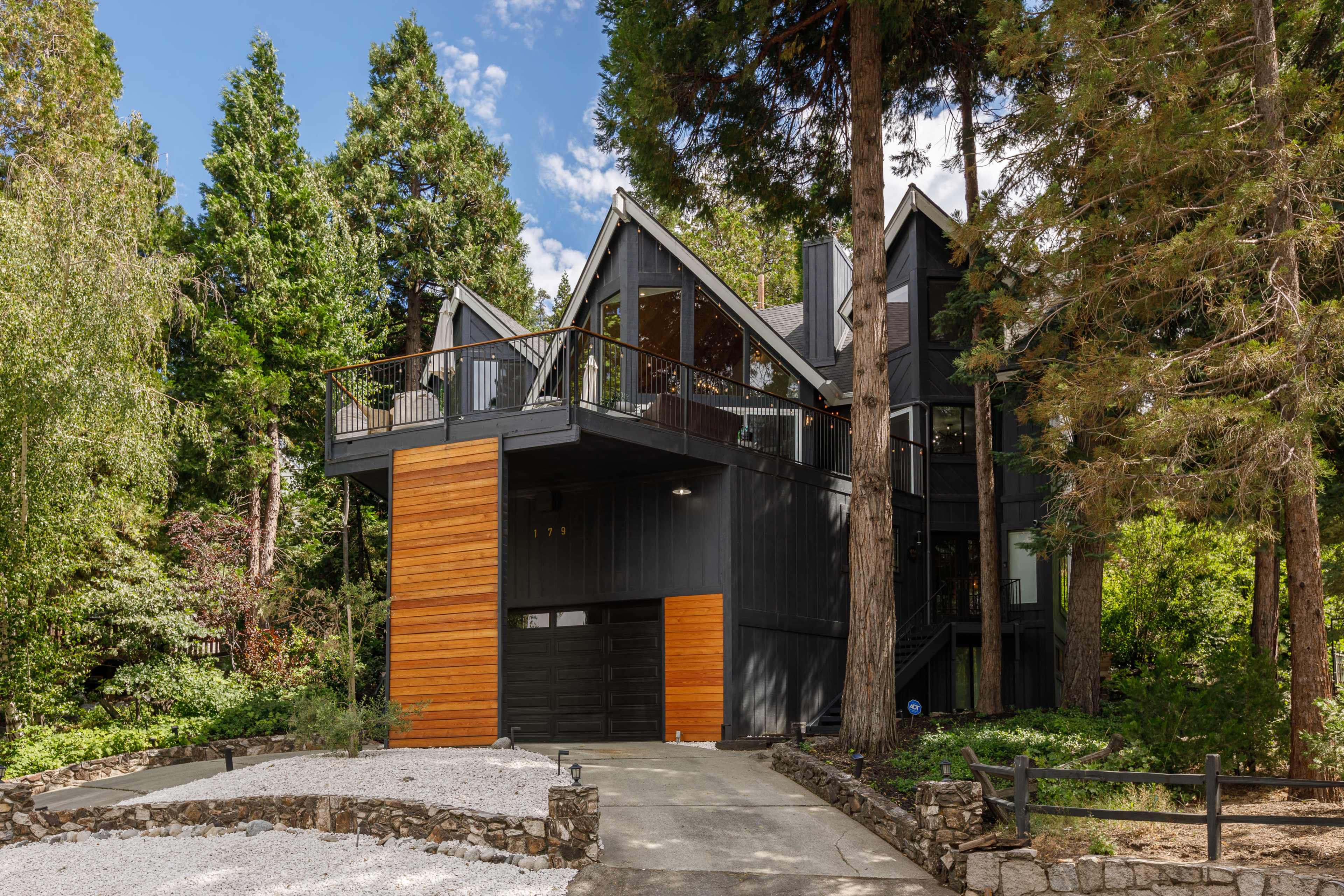 A modern black and wood house set among tall trees, featuring a garage and a balcony overlooking the surrounding landscape.
