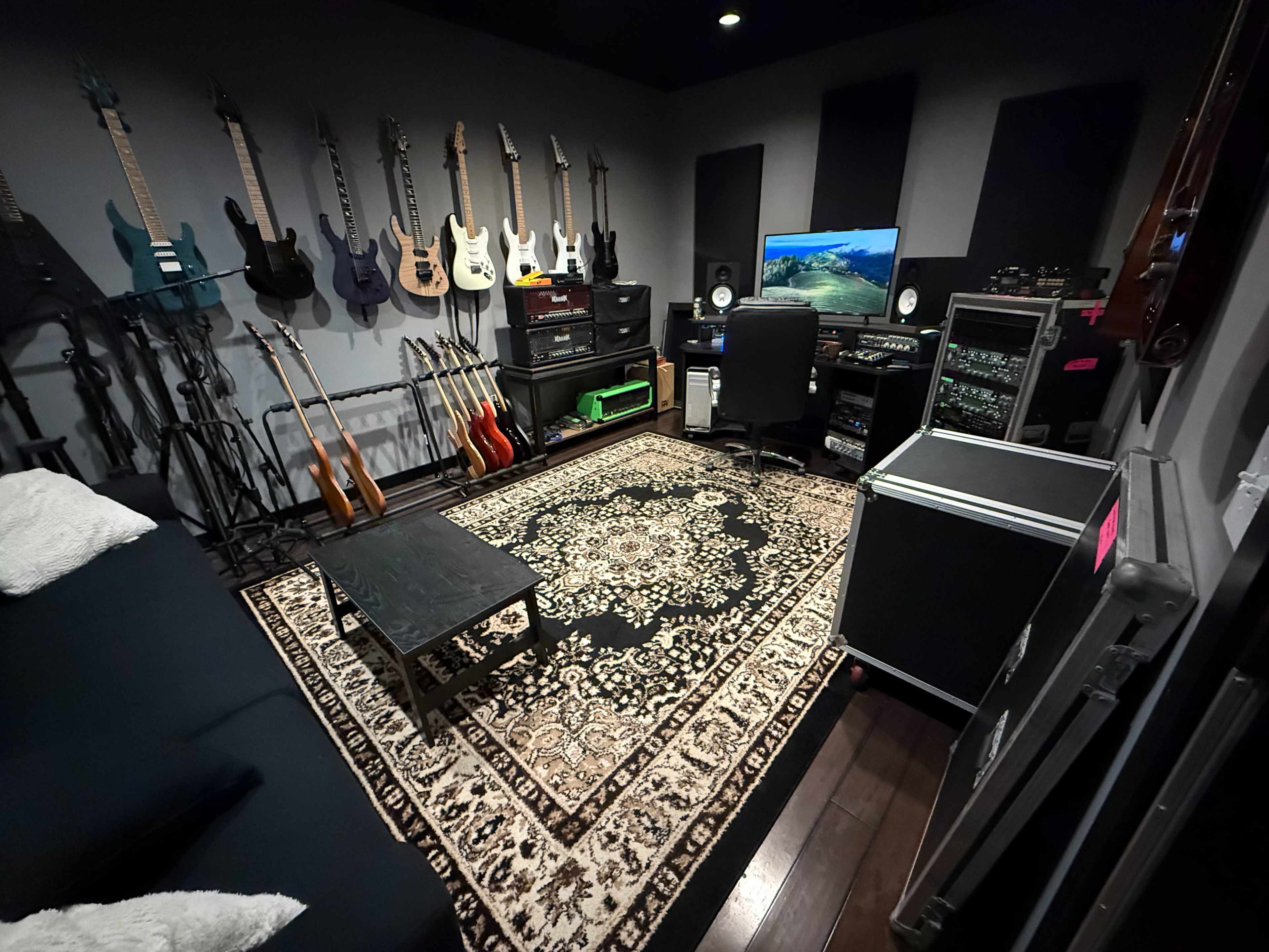 The image shows a music recording studio with various guitars hanging on the wall, a computer setup, and a patterned rug on the floor.