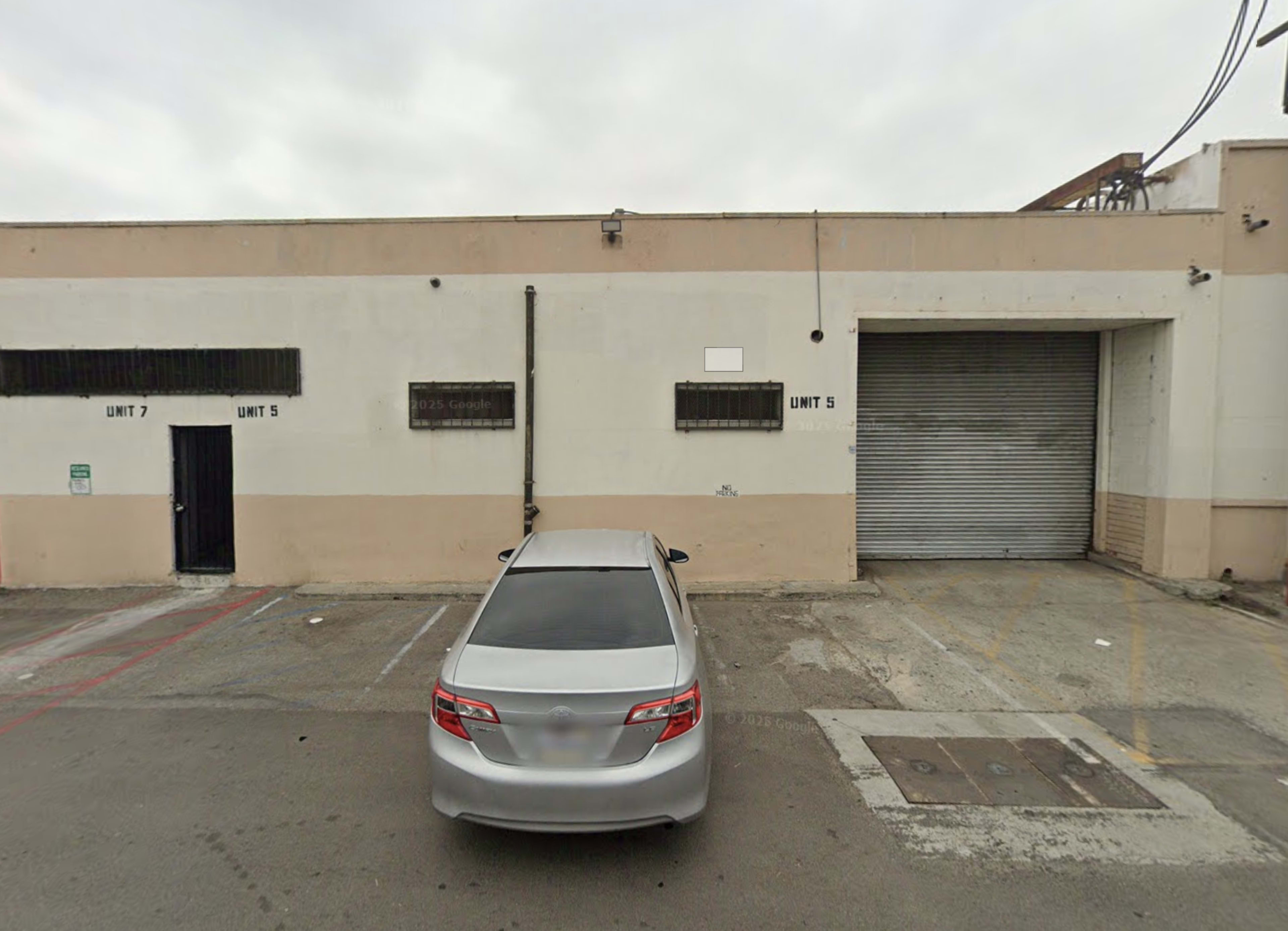 The image shows a gray car parked in front of a warehouse with the numbers "UNIT 5" and "UNIT 7" visible on the building.