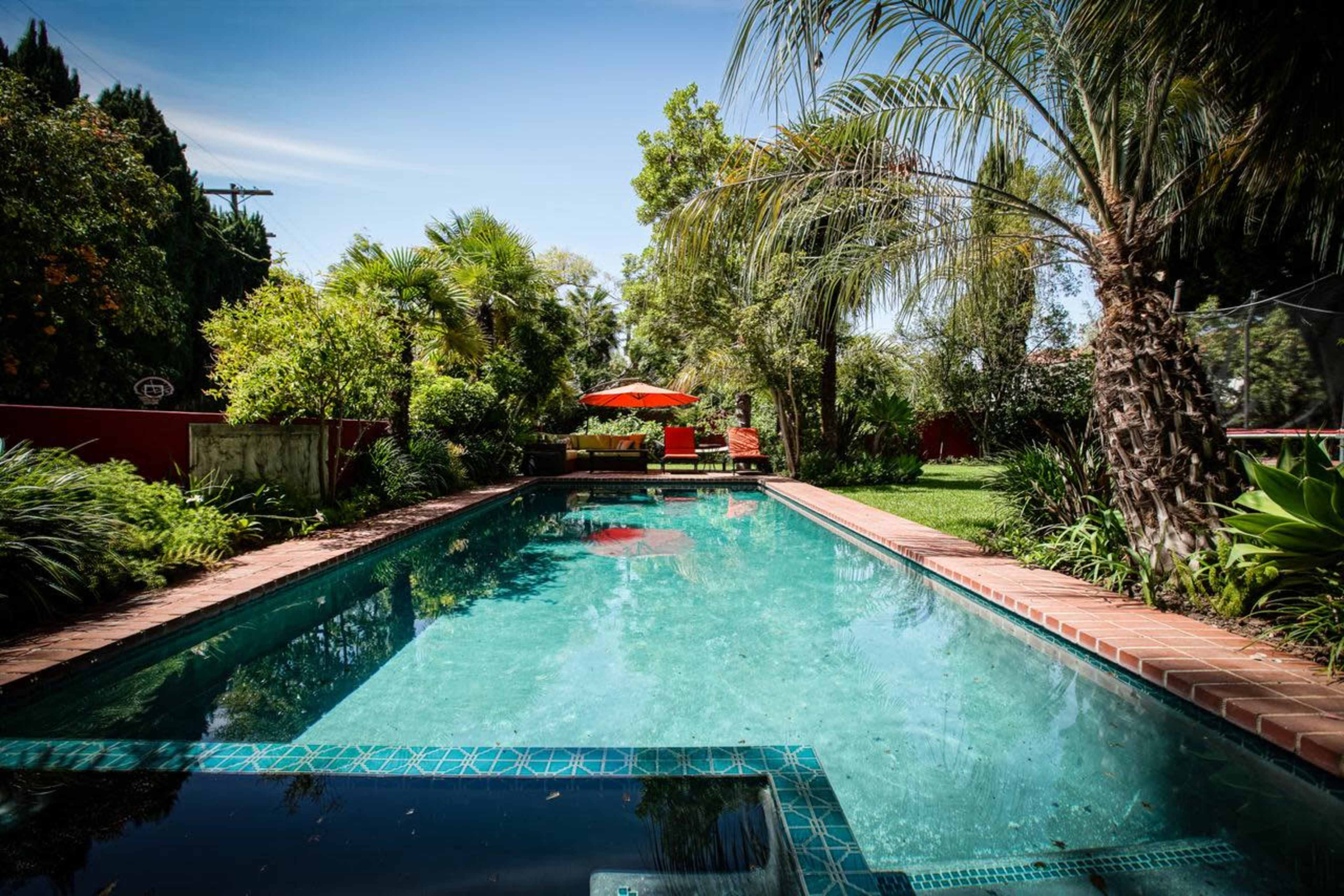 A clear swimming pool is framed by lush greenery and a shaded seating area under an orange umbrella.