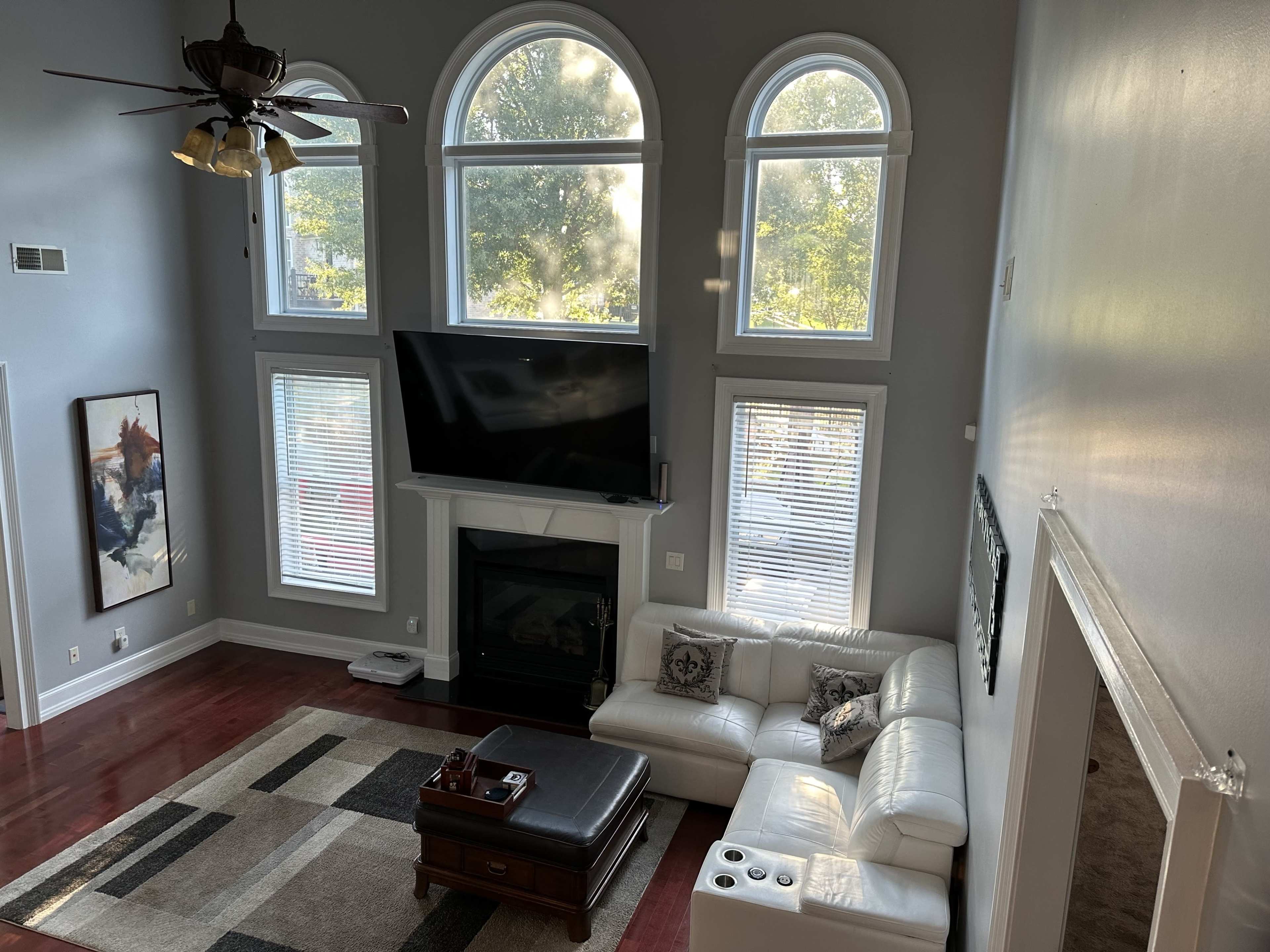 The image depicts a modern living room with large arched windows, a flat-screen TV mounted above a fireplace, a white sectional sofa, and a coffee table on a patterned rug.