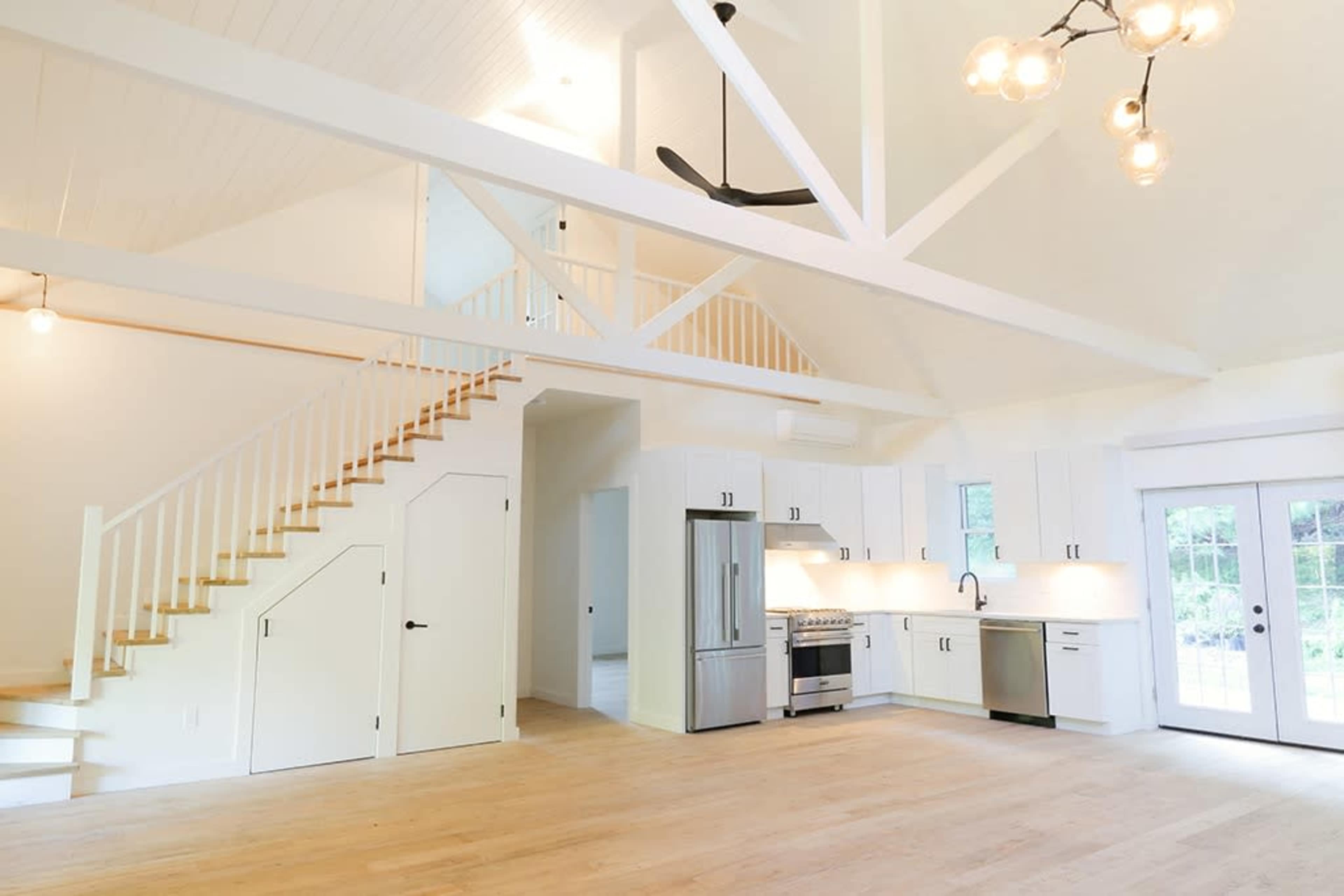 A bright, open-concept living area with a modern kitchen, a staircase leading to a loft, and large windows that allow natural light to enter.