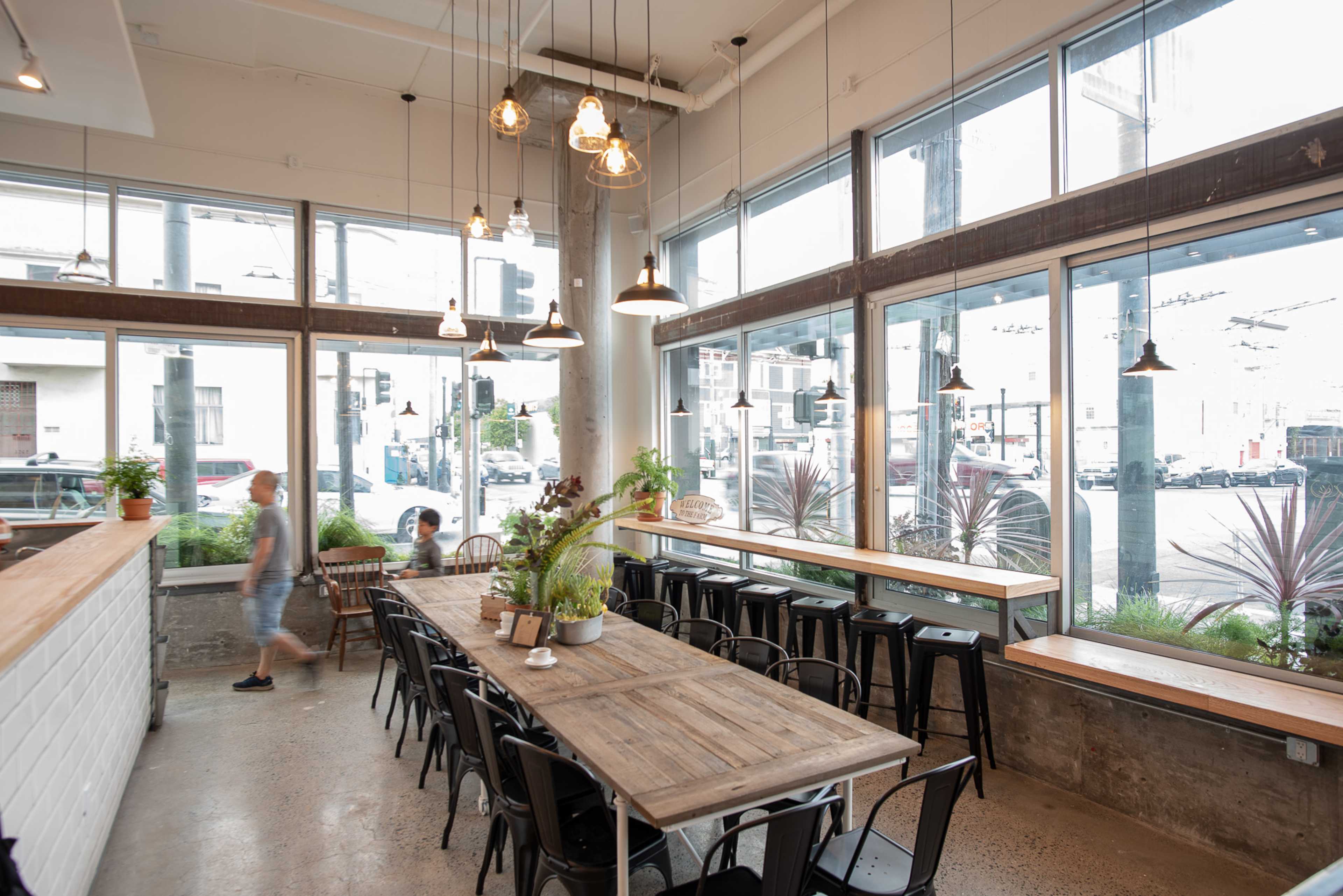 A spacious cafe features a large wooden table surrounded by black chairs, with large windows allowing natural light to fill the room.