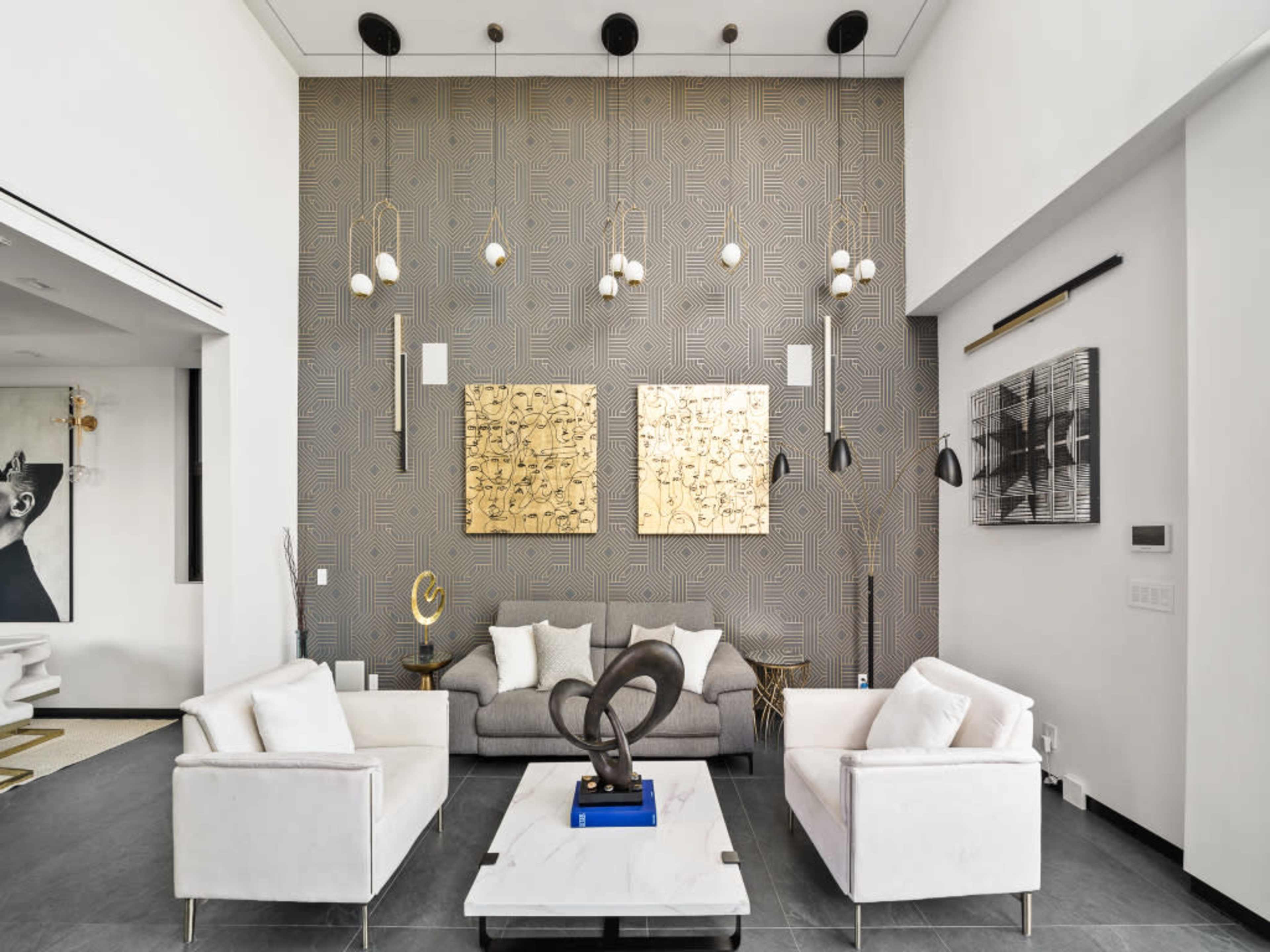 The image shows a modern living room with a high ceiling, featuring a gray sofa, two white armchairs, contemporary art on the walls, and decorative pendant lights.