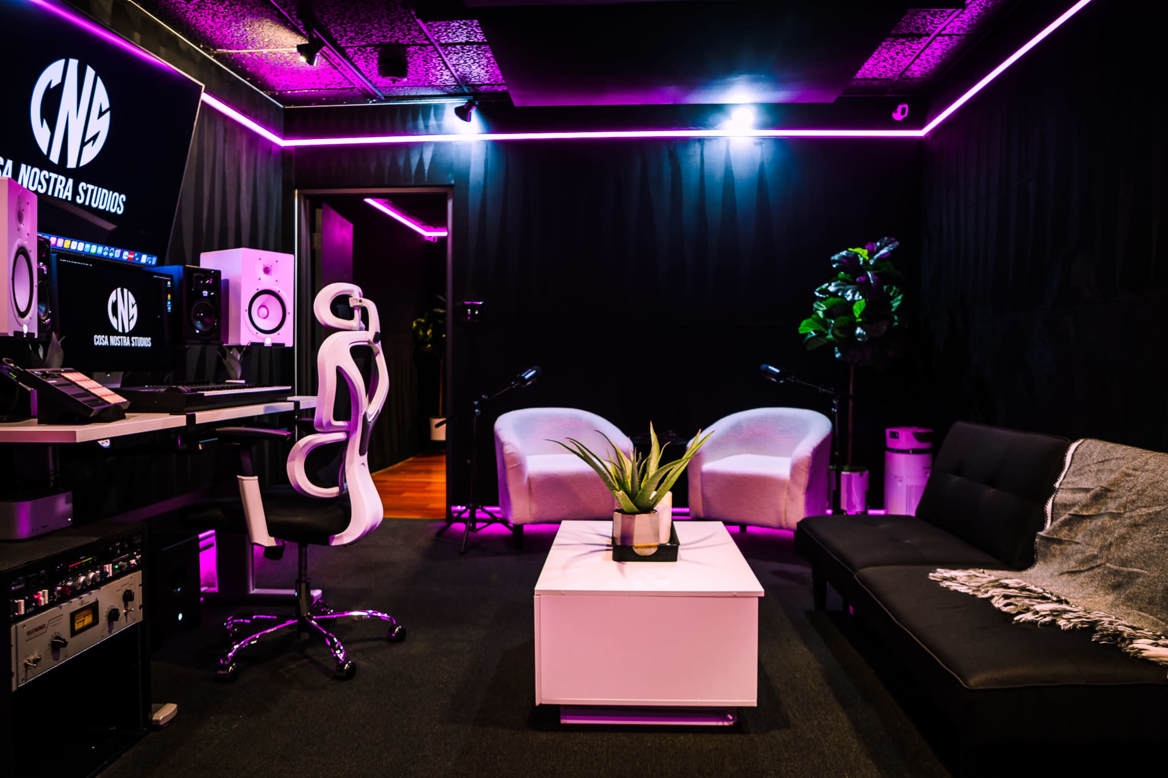 The image shows a modern studio space with a black sofa, two white chairs, a table, and vibrant LED lighting.