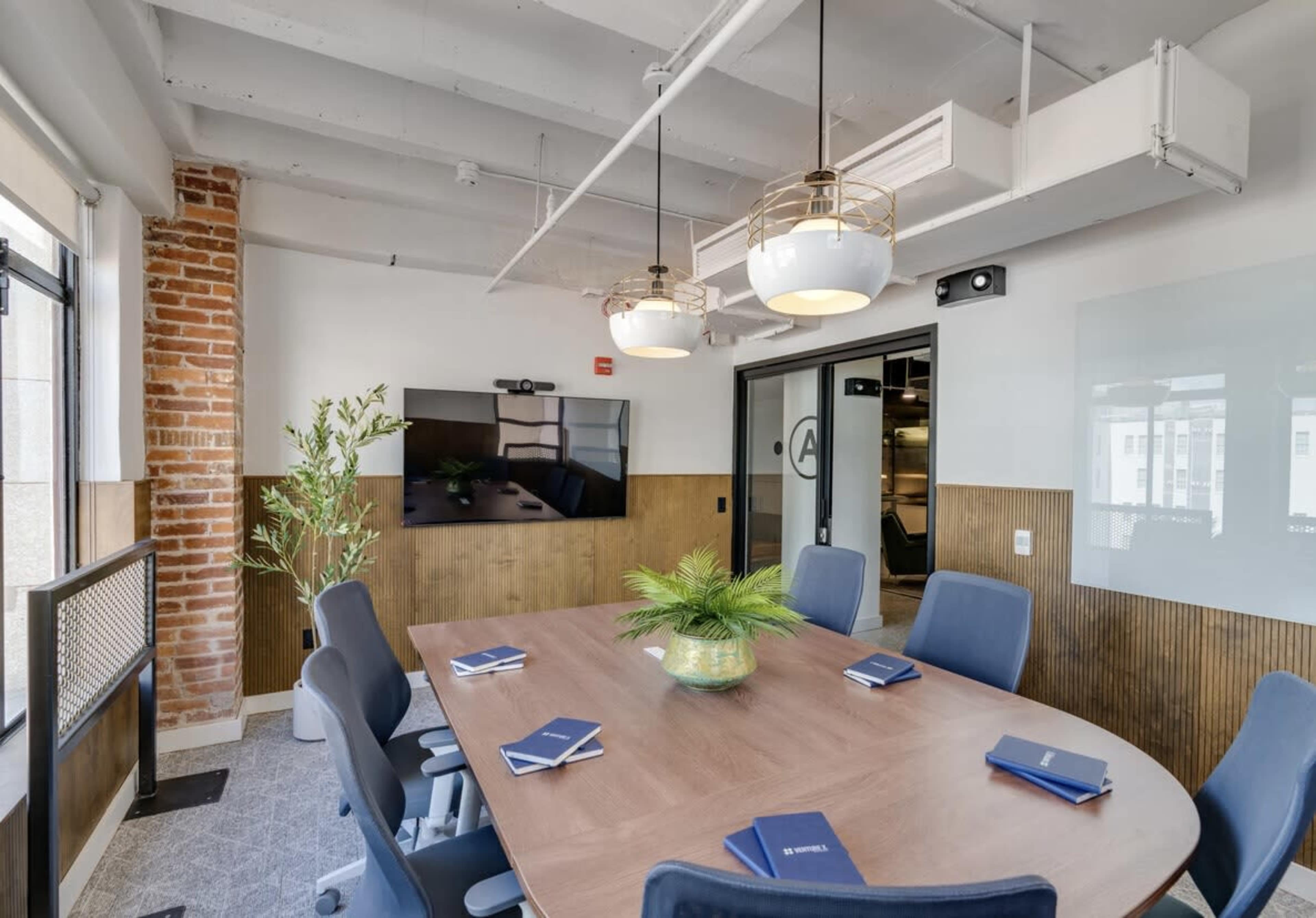 A modern conference room features a large oval table surrounded by blue chairs, with a television screen on one wall and decorative plants.