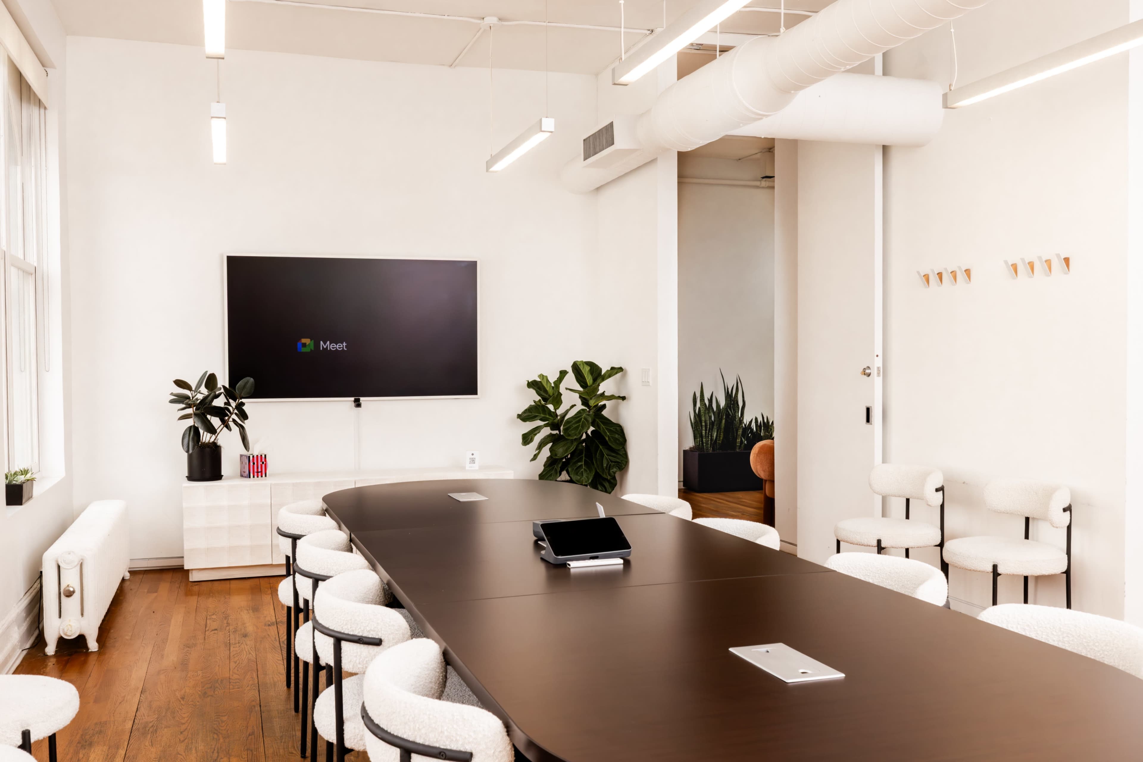 A modern conference room features a long dark table surrounded by white chairs, a television mounted on the wall, and potted plants in the corners.