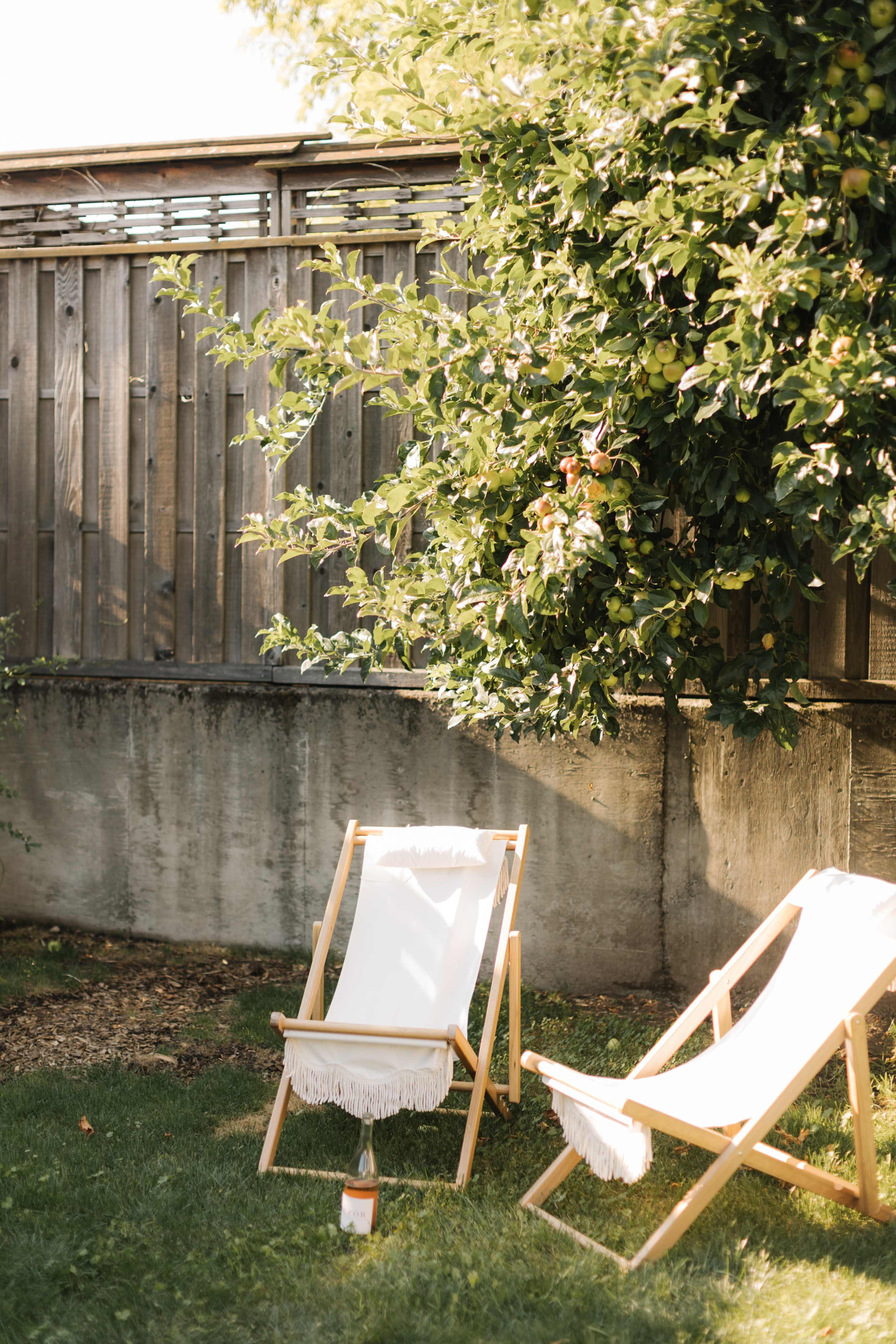 Two wooden lounge chairs with white fabric are placed on grassy ground beside a wooden fence and a tree.
