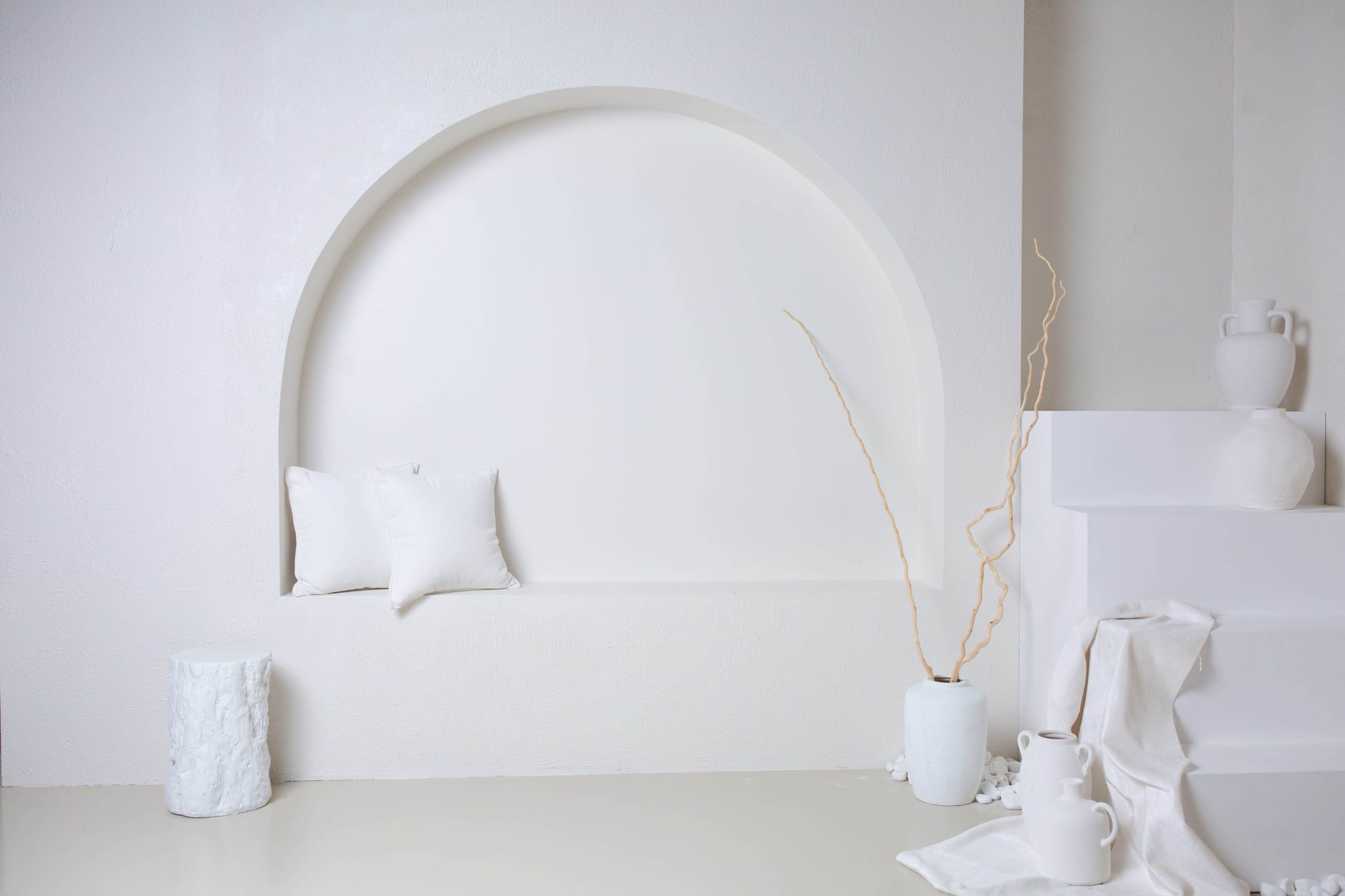 The image shows a minimalistic interior with a white curved niche featuring two pillows, a decorative twig in a vase, and a textured cylindrical pedestal.