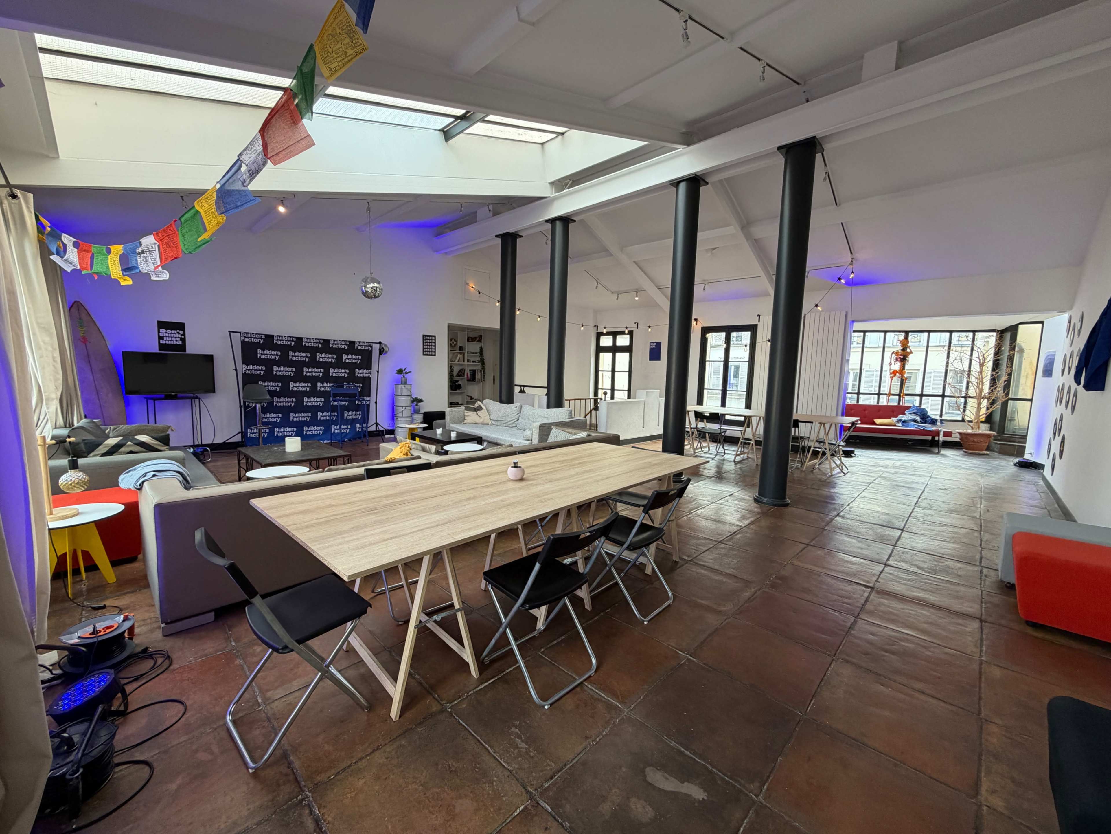 The image shows a spacious, modern communal area with seating arrangements, a large table, and colorful decorations hanging from the ceiling.