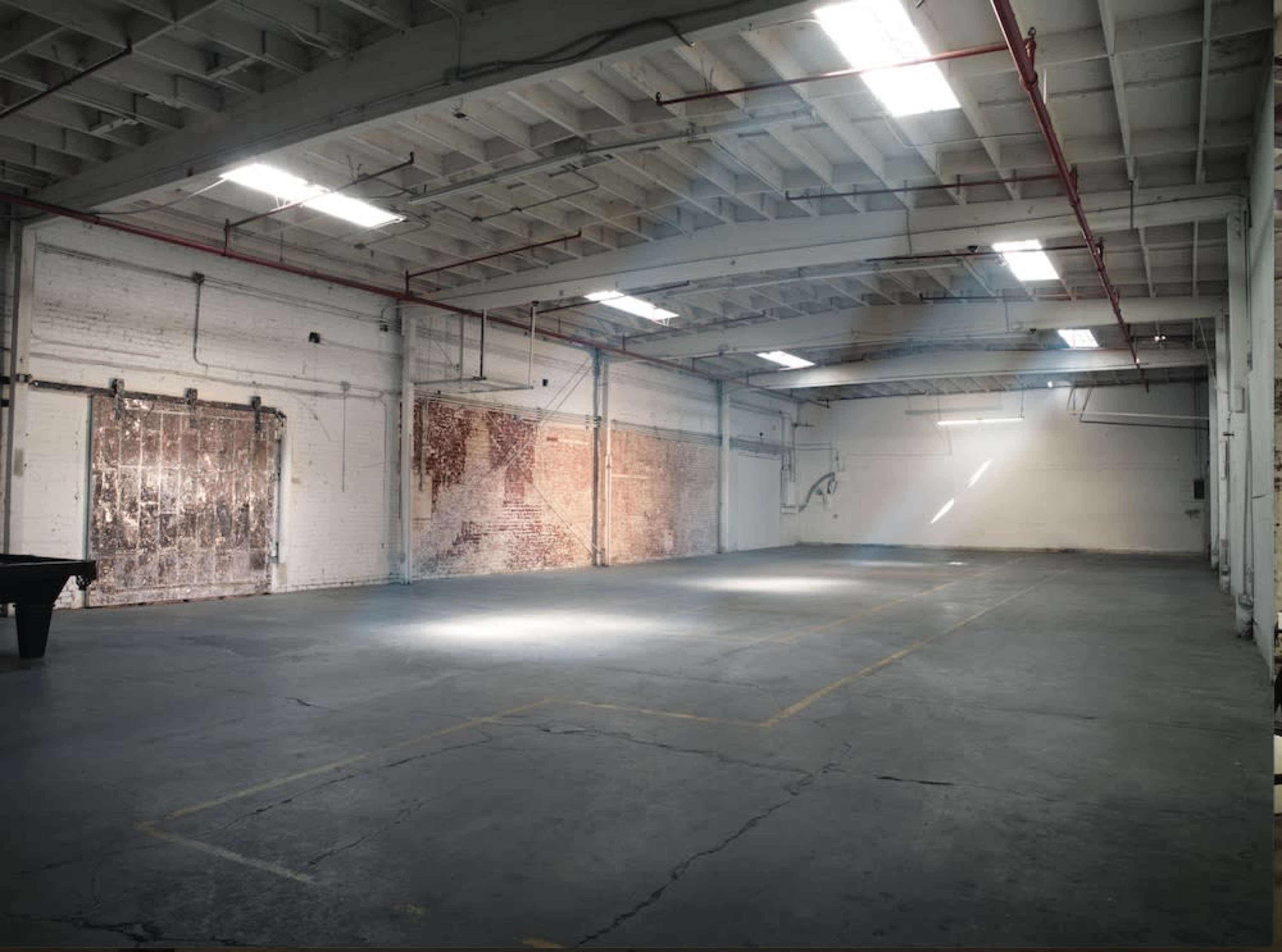 An empty industrial warehouse features exposed brick walls and sunlight streaming through the skylights, illuminating the concrete floor.