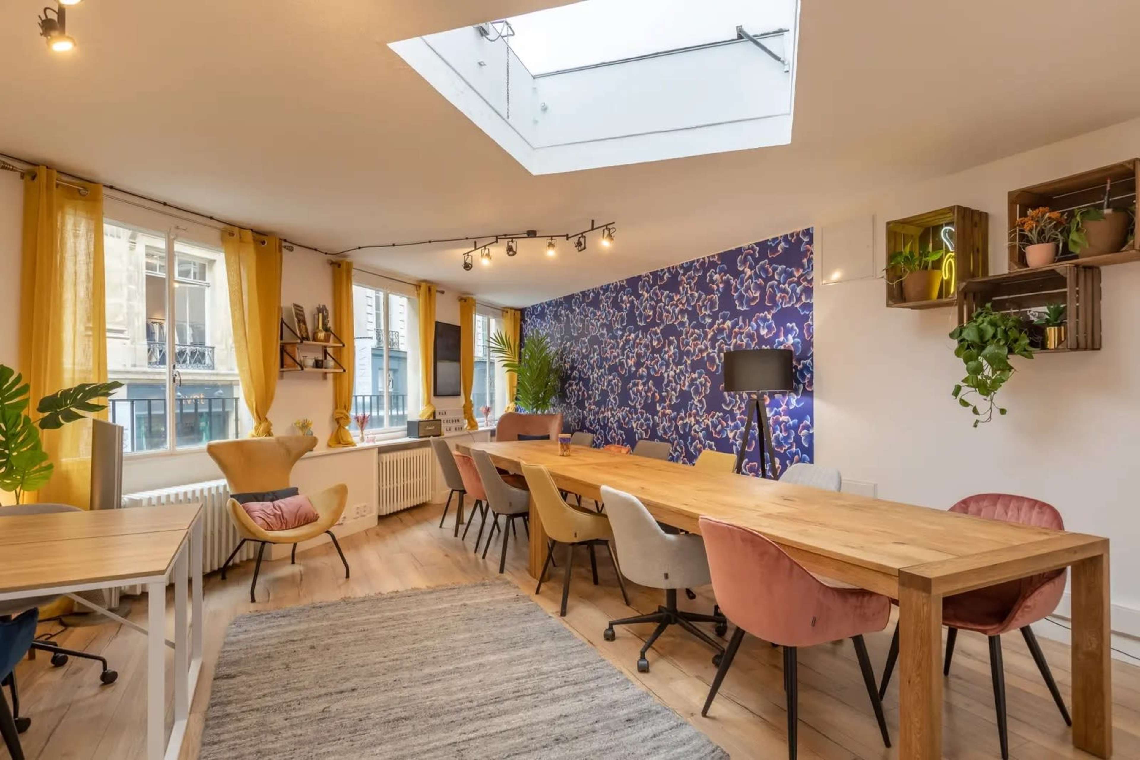 The image shows a modern office space with wooden tables, colorful chairs, and large windows illuminated by natural light.