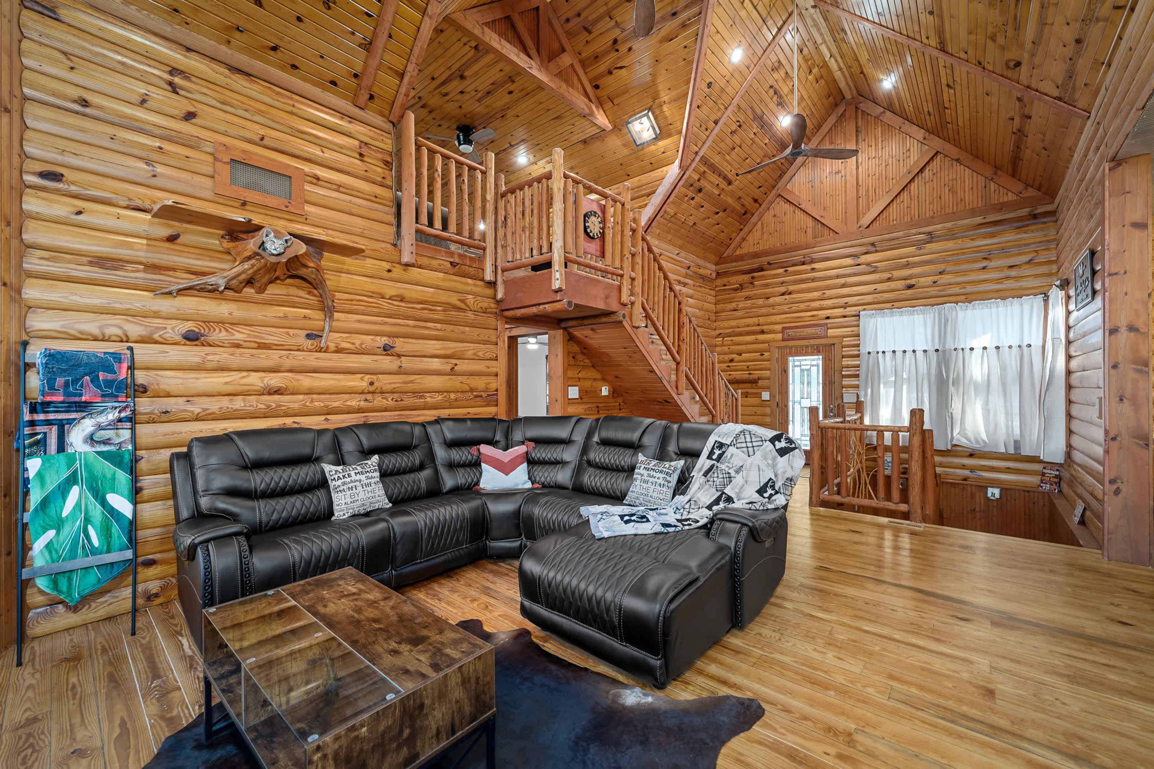A spacious wooden living room featuring a large black sectional sofa, a glass coffee table, and a wooden staircase leading to an upper loft area.