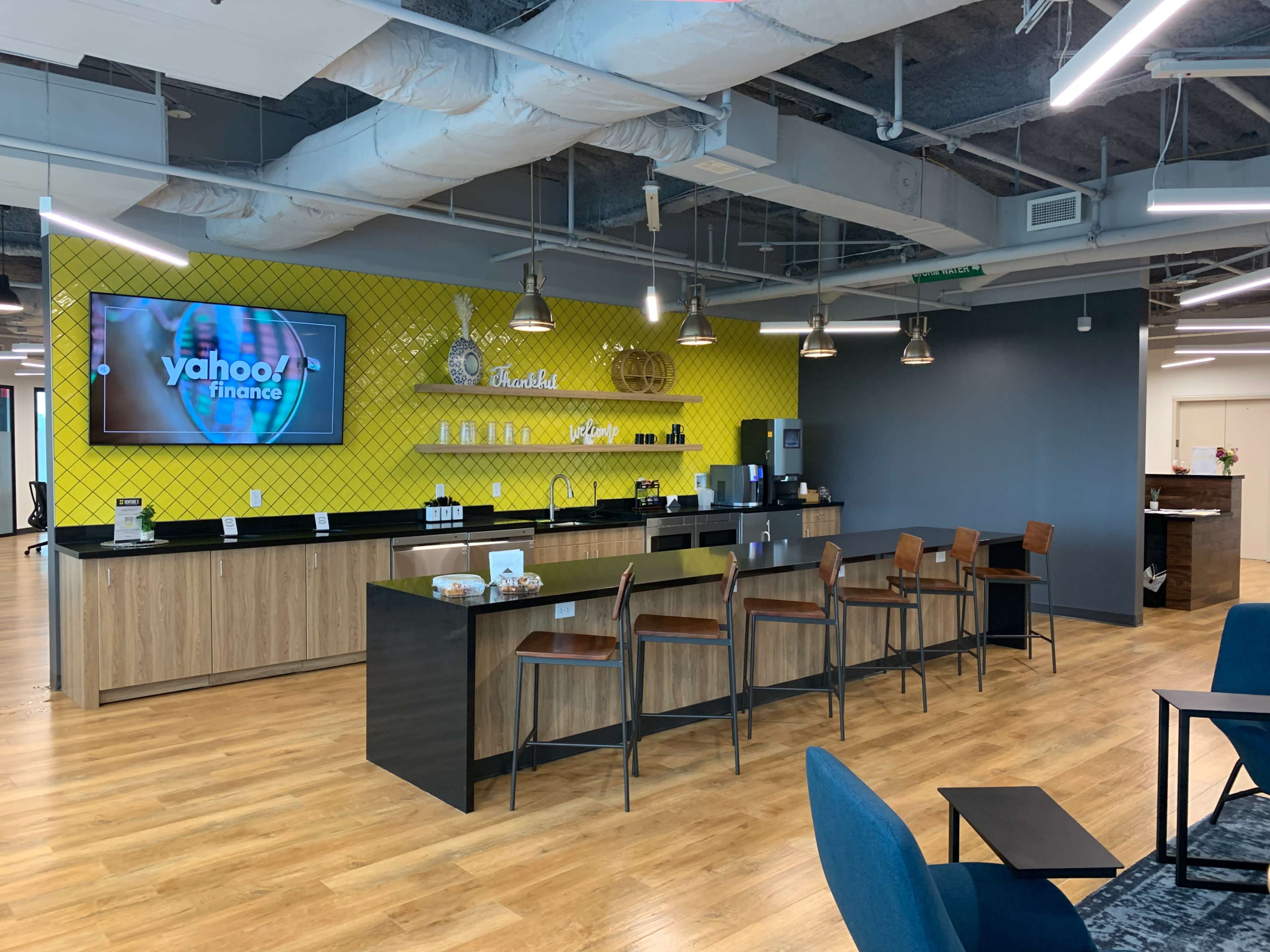 The image shows a modern office kitchenette area with a long bar featuring high stools, a large screen displaying "Yahoo Finance," and various kitchen amenities against a bright yellow patterned wall.