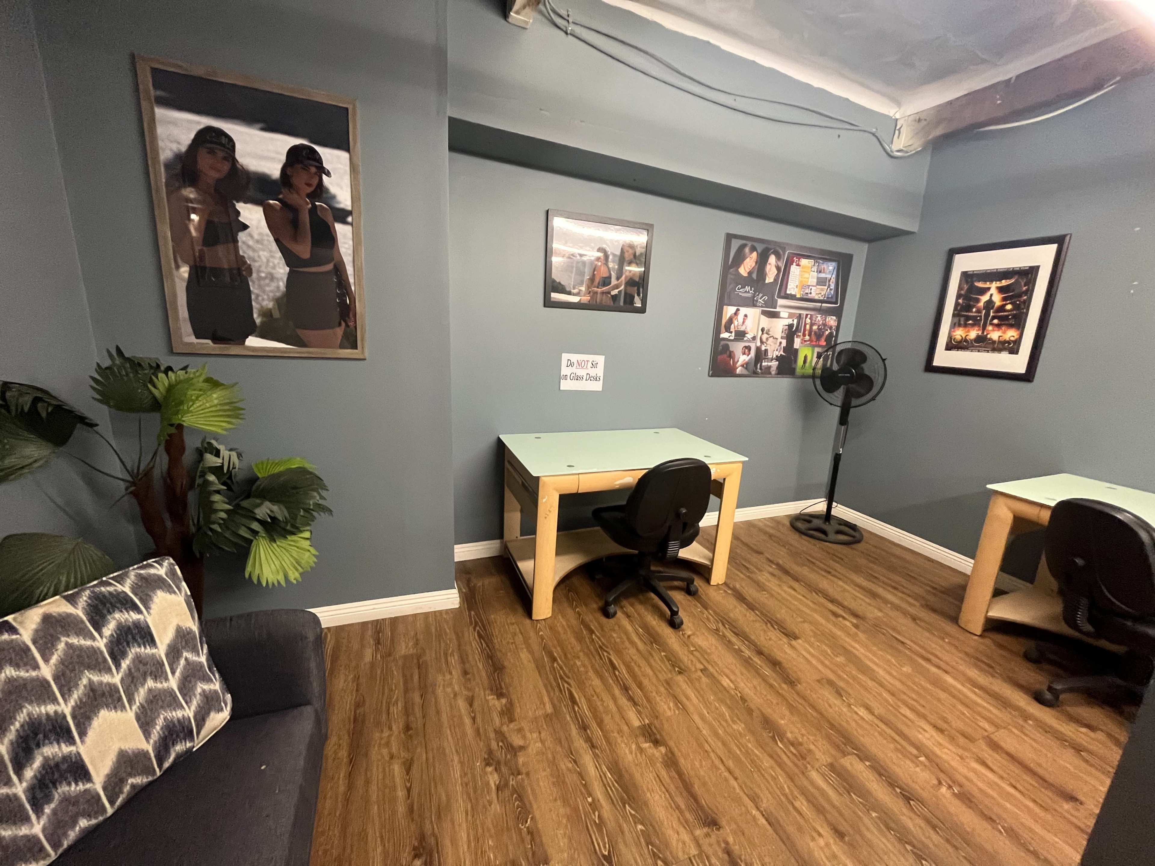 The image shows a room with two desks, a couch, framed photographs on the walls, and a fan in the corner.