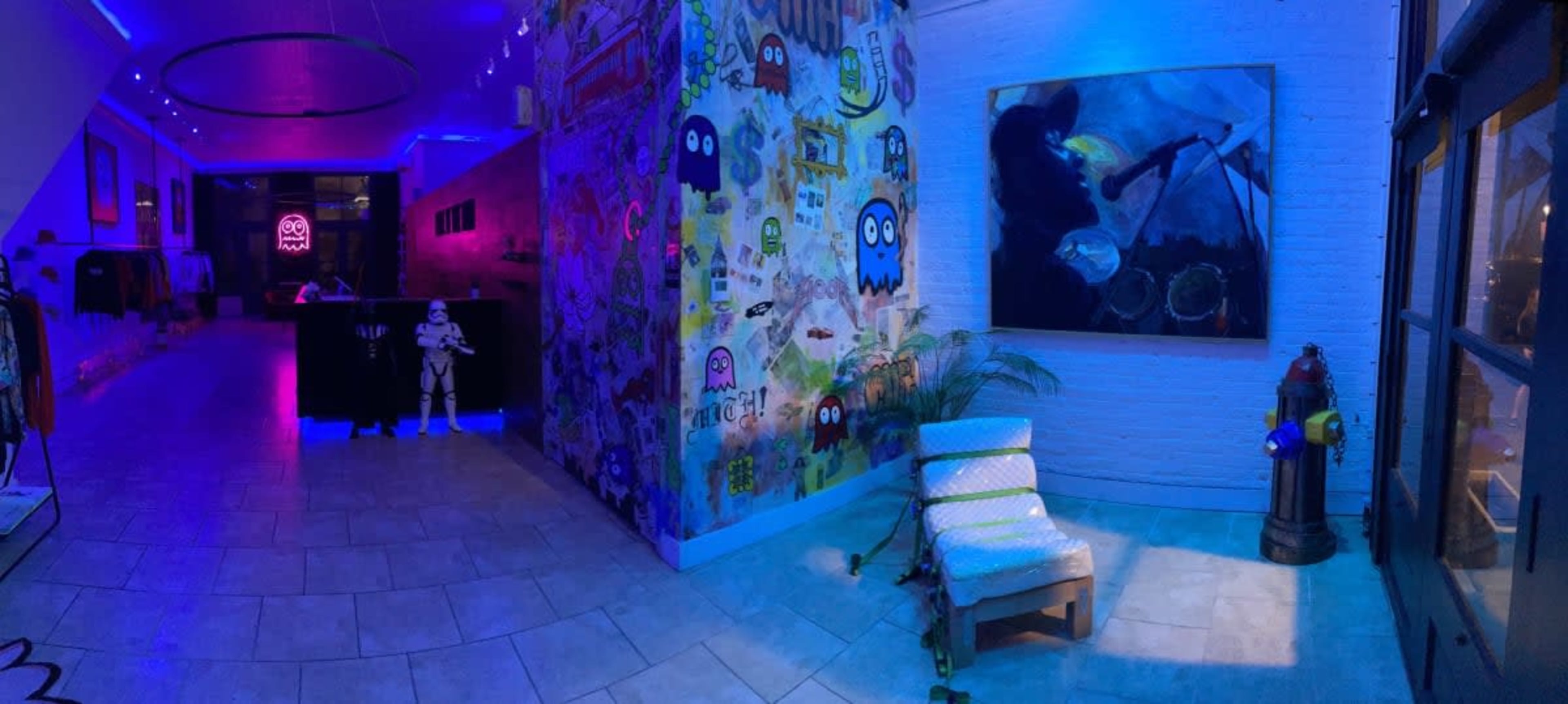A brightly lit interior space featuring graffiti-covered walls, a lounge chair, a fire hydrant, and a life-sized stormtrooper figure under blue and purple lighting.