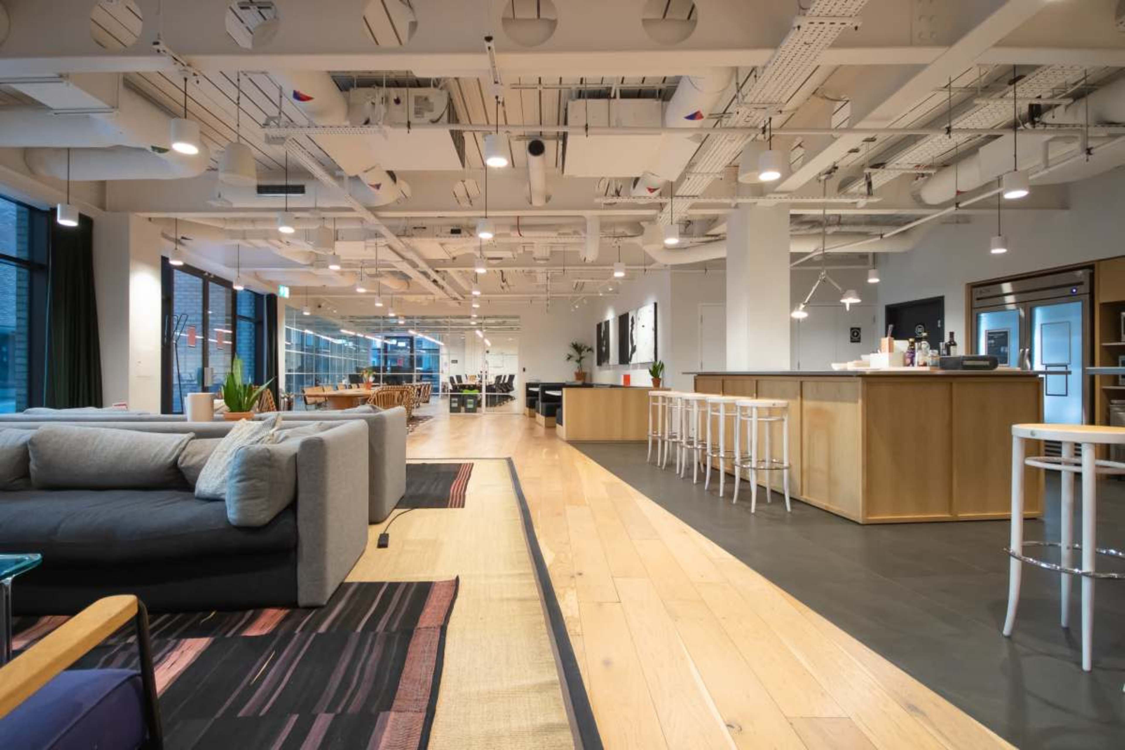 The image shows a modern coworking space with a combination of seating areas, a bar counter, and large windows allowing natural light to fill the room.