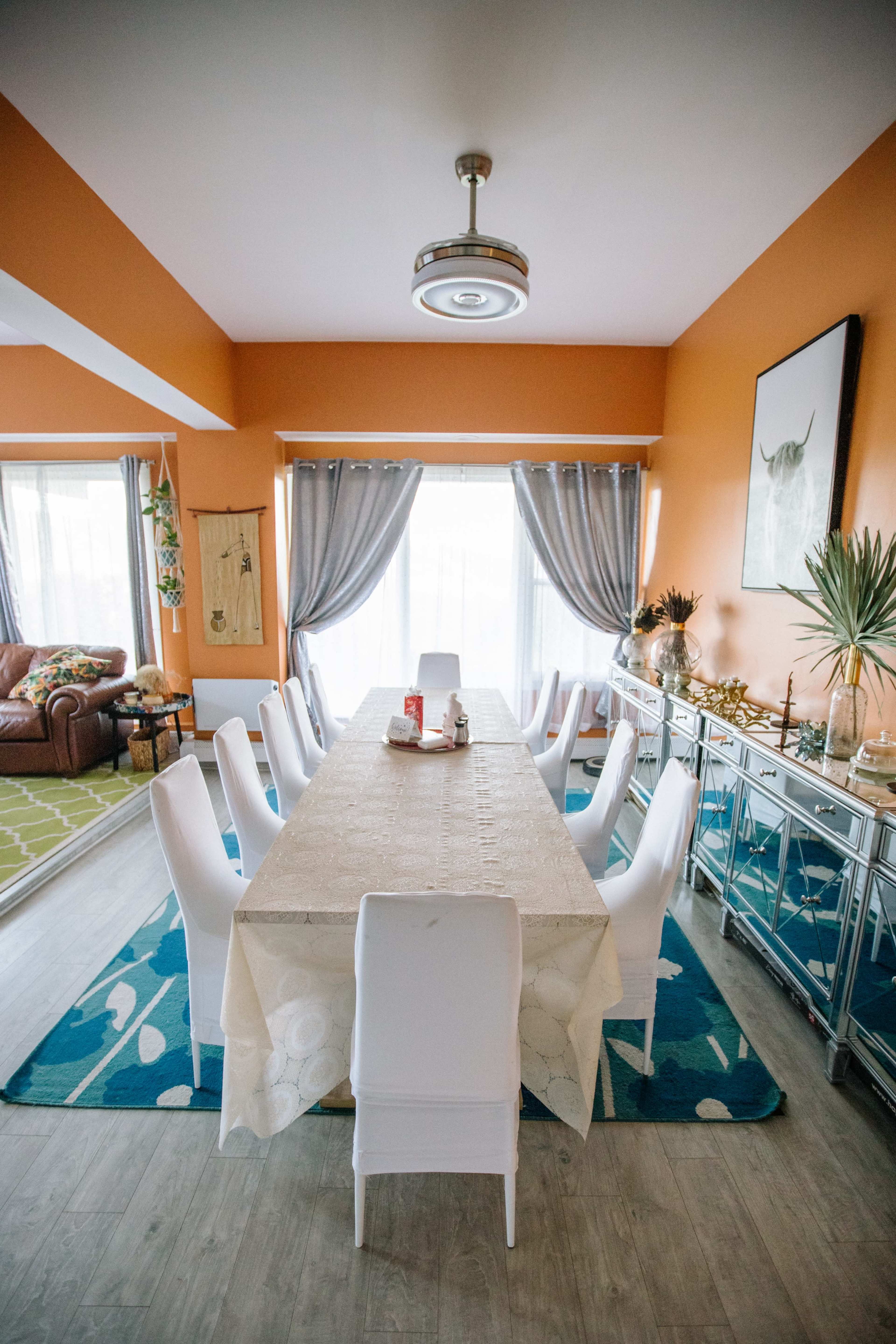 A long dining table is set in a brightly colored room with orange walls and large windows, surrounded by white chairs and a blue area rug.