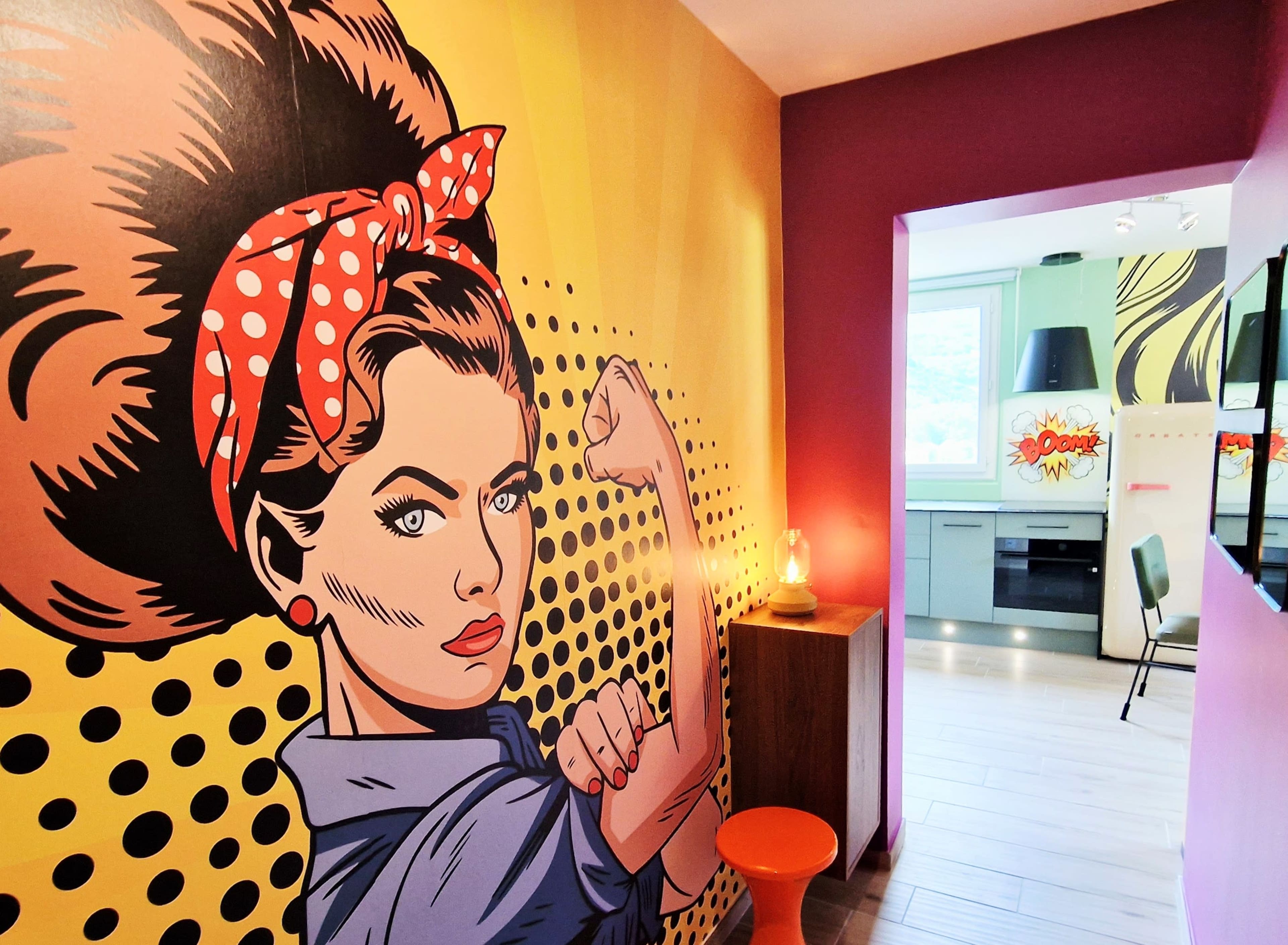 The image shows a brightly colored hallway featuring a large pop art mural of a woman with a polka dot headband and a bold pose, leading to a kitchen area.
