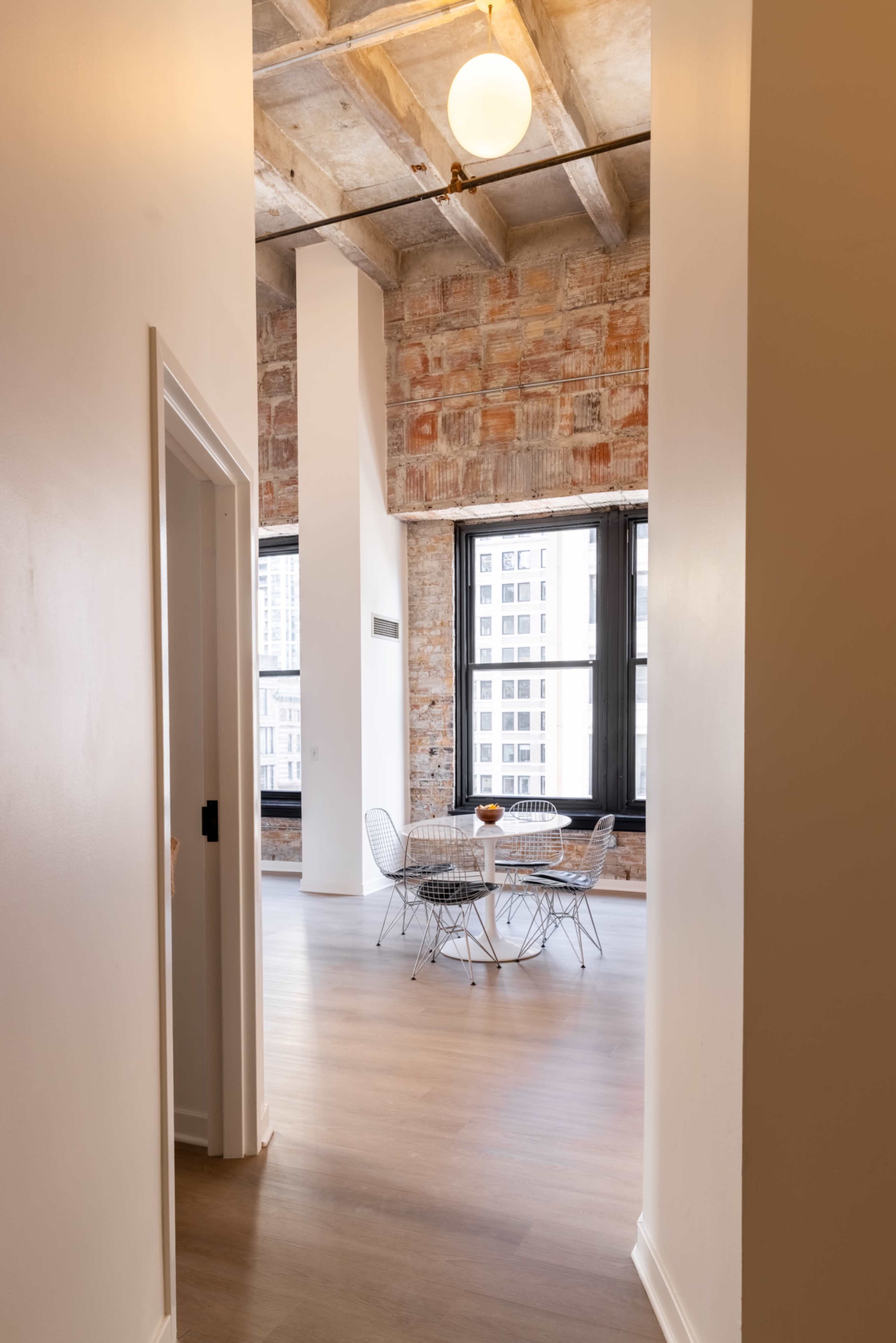 A hallway leads to a dining area with a round table and wire chairs, framed by exposed brick walls and large windows.