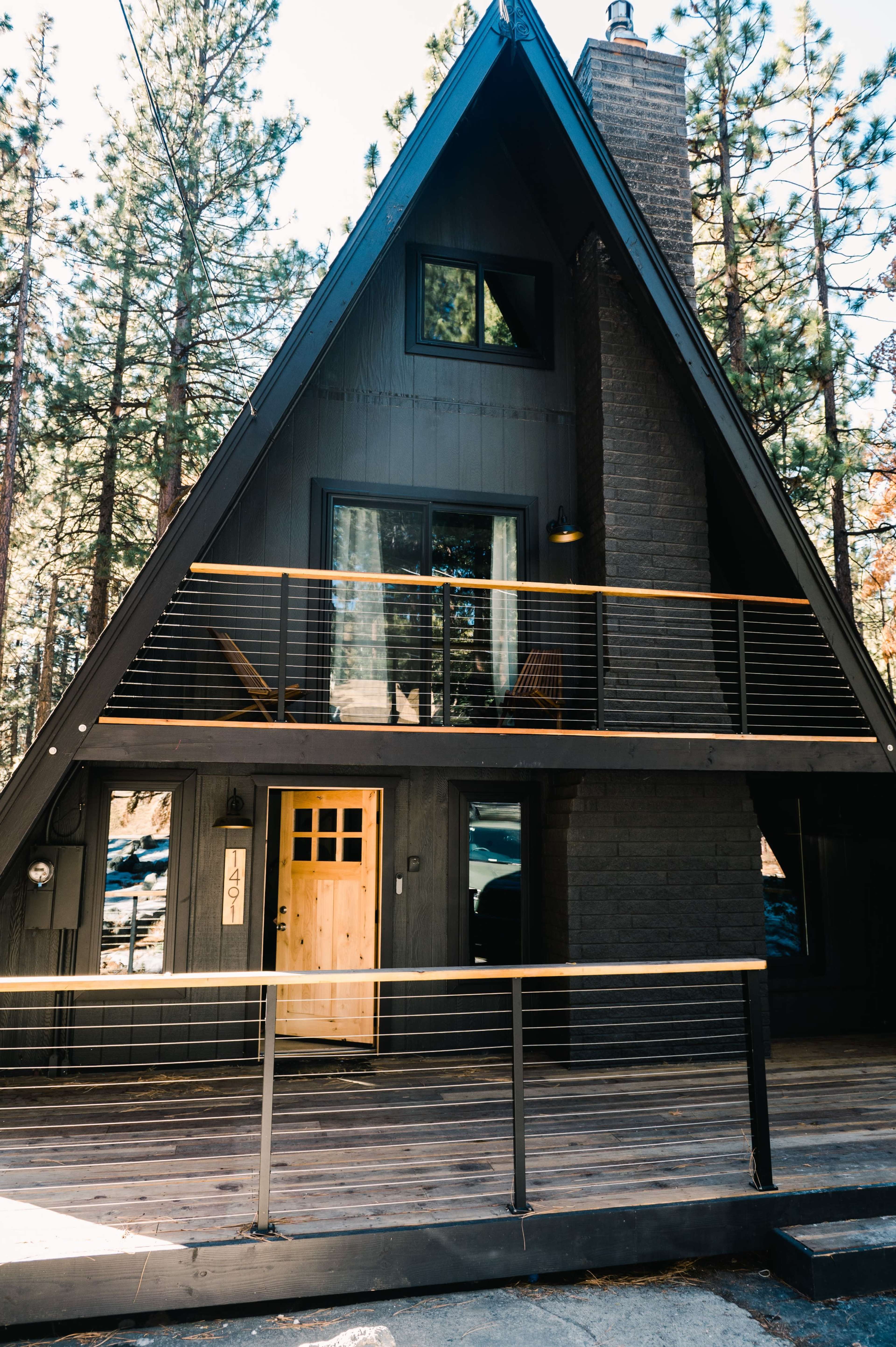 A modern A-frame house with large windows and a wooden entrance, situated among tall trees.