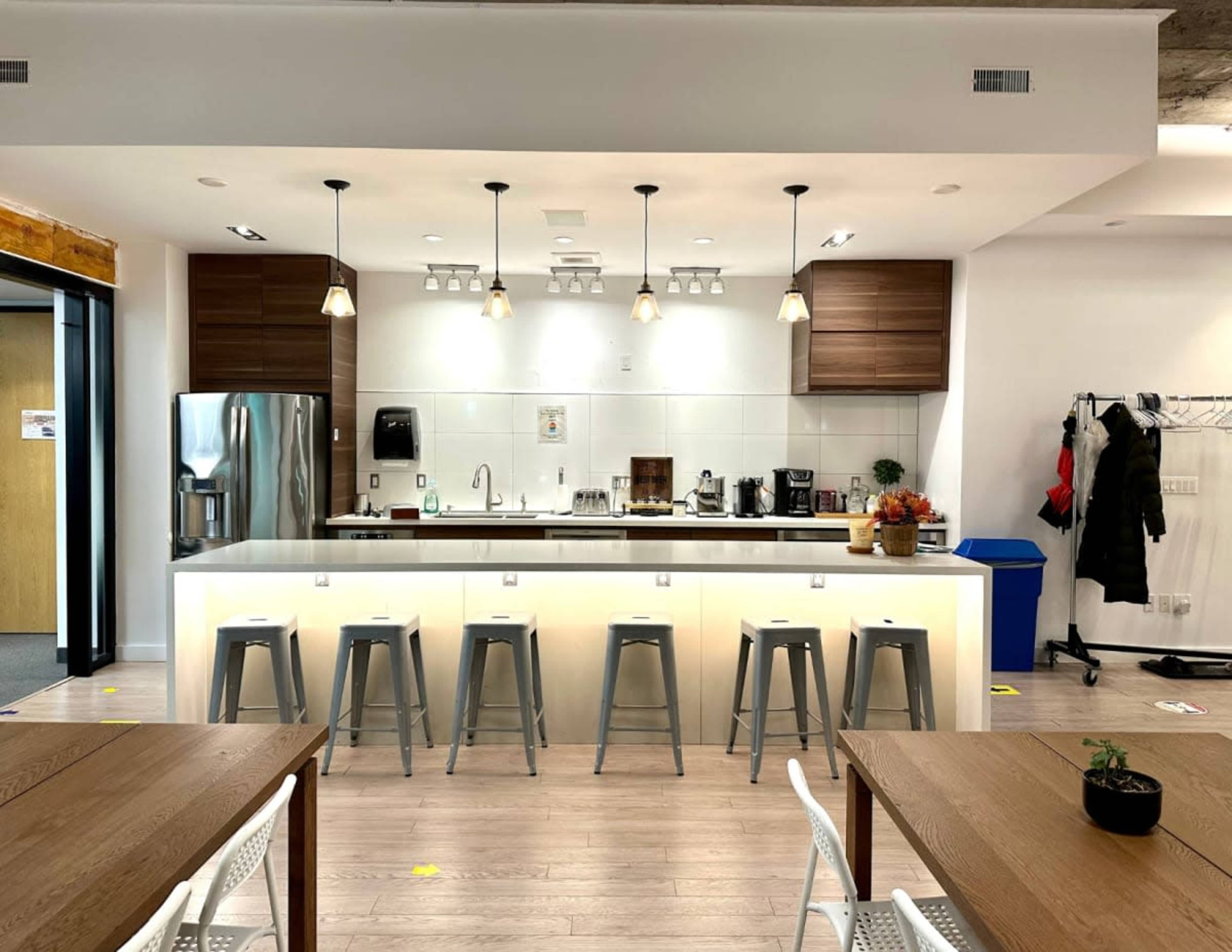 The image shows a modern kitchen area with a countertop, bar stools, and various kitchen appliances, including a refrigerator and coffee maker.