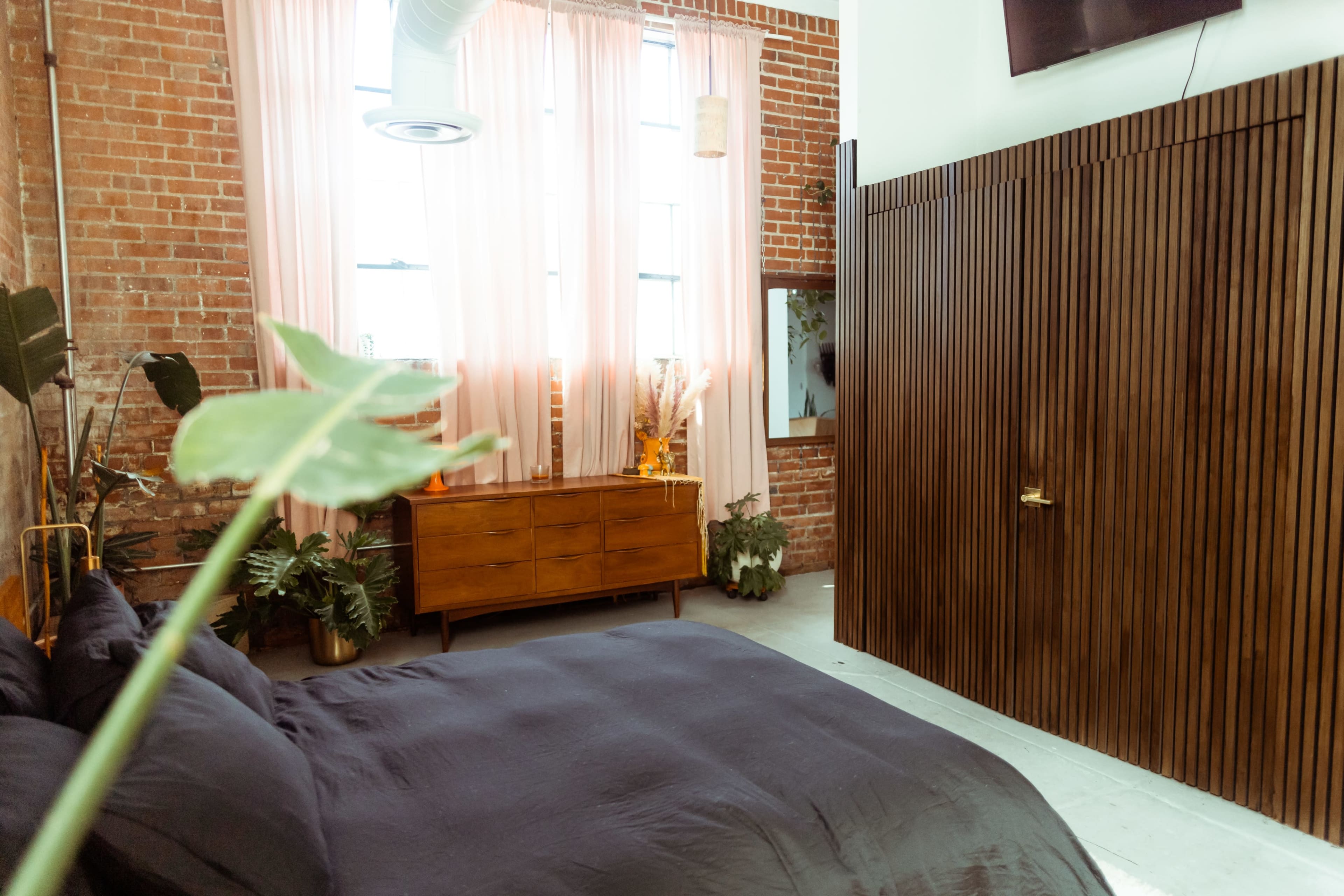 A bedroom with a brick wall, large windows covered by pink curtains, a wooden dresser, and a dark bedspread.