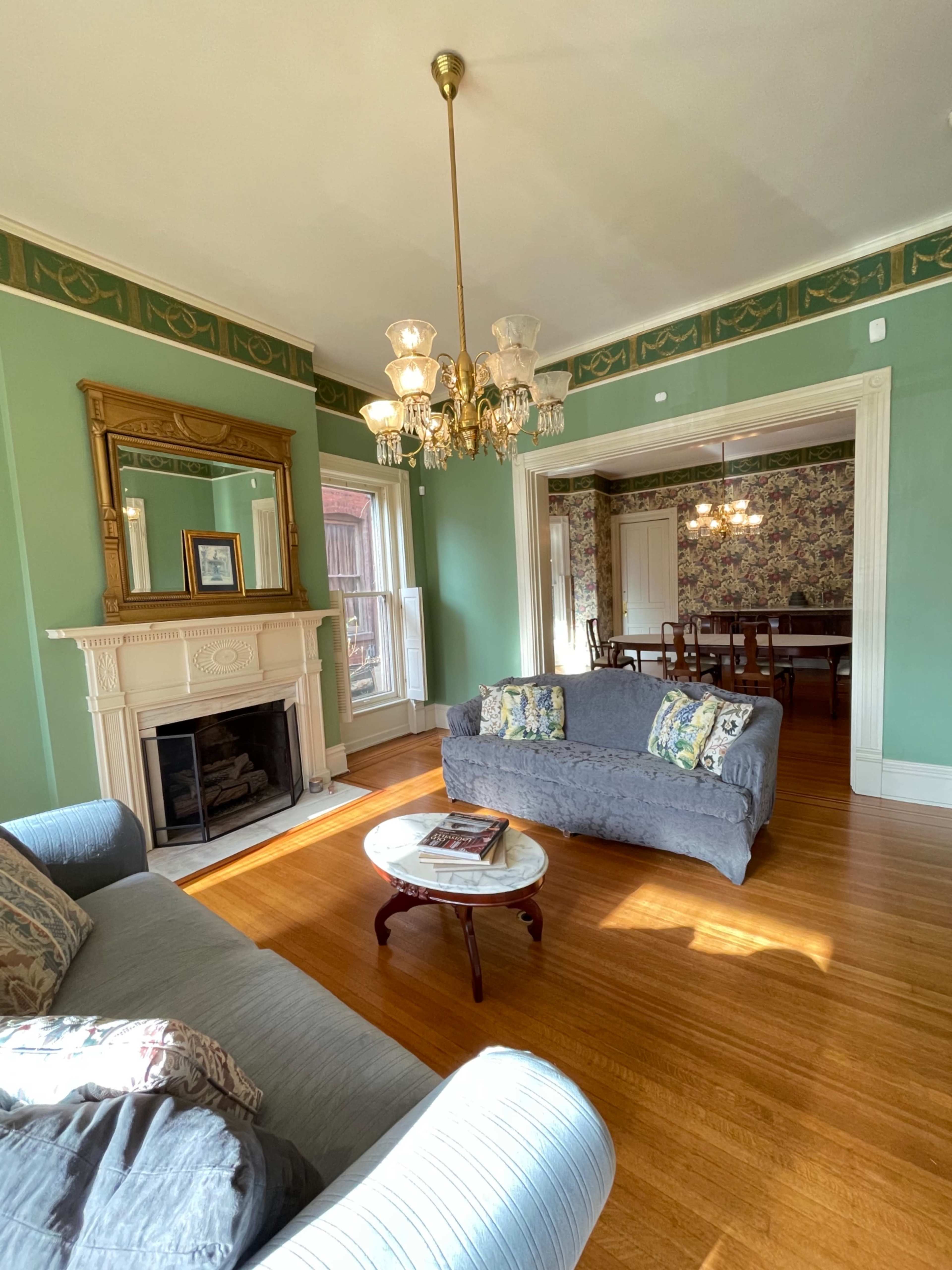 The room features a light green wall with a chandelier, a fireplace with a mirror above it, and a cozy seating area with two sofas and a round table.