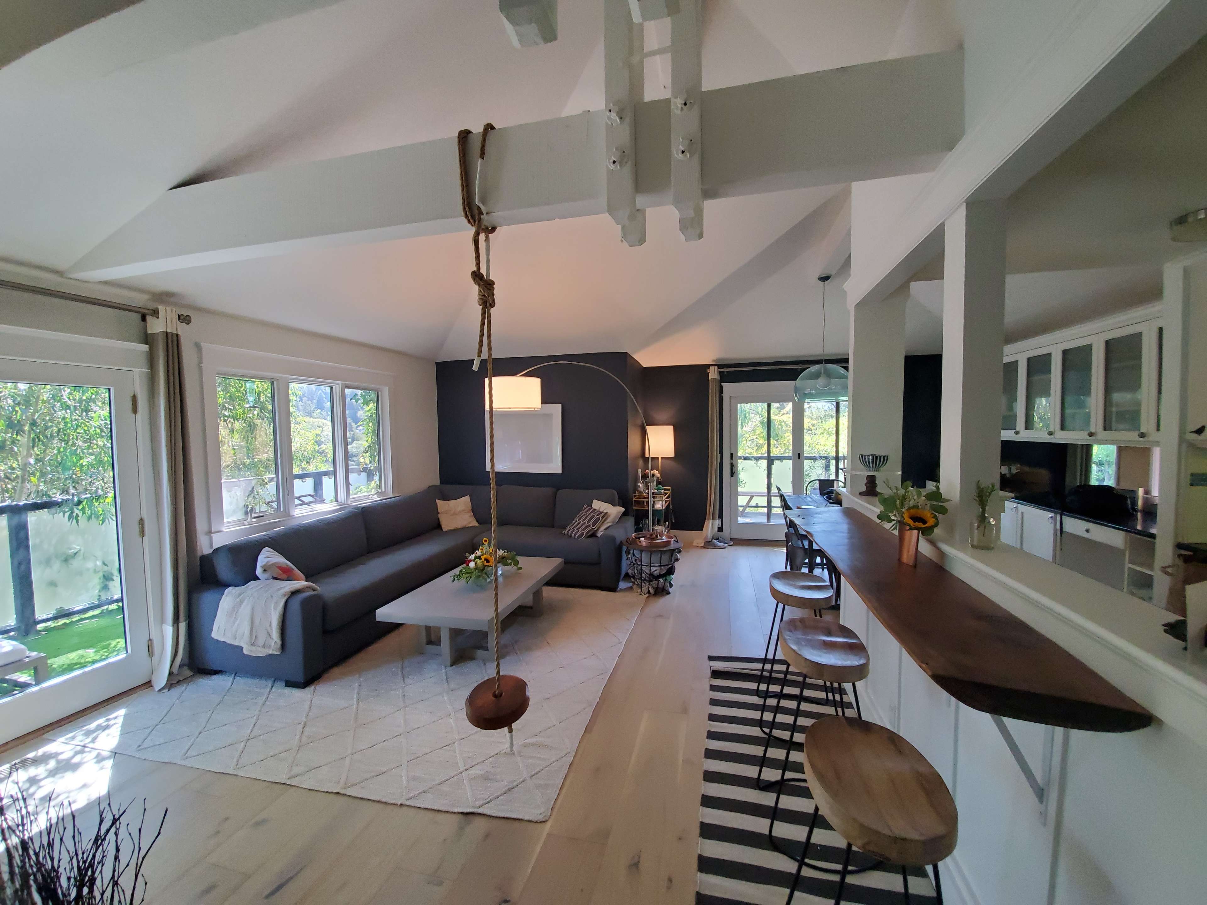 A modern living room features a gray sectional sofa, a wooden bar with stools, and a swing hanging from the ceiling, surrounded by large windows that provide natural light.
