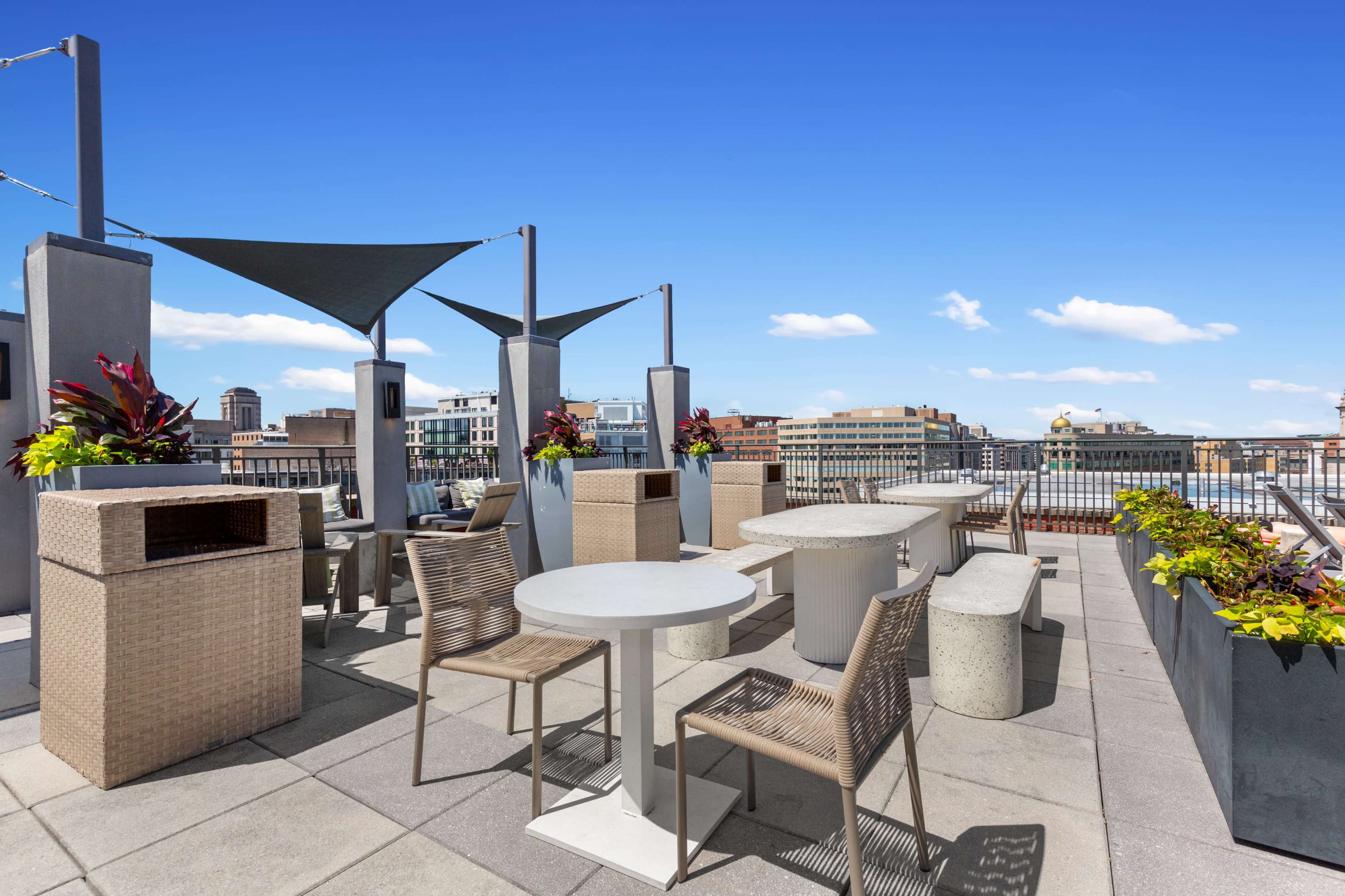 The image shows a rooftop terrace featuring several tables and chairs surrounded by planters with colorful plants against a clear blue sky.