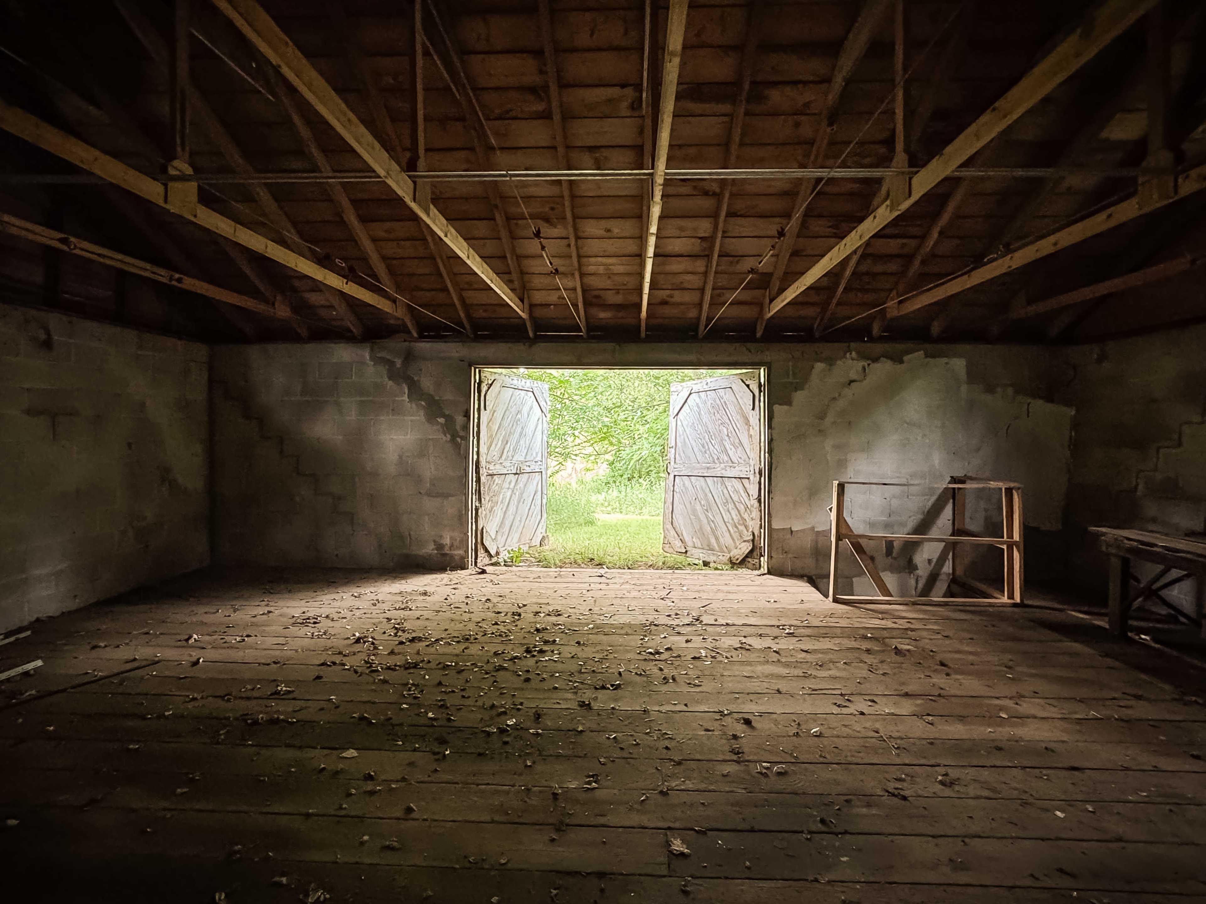 An empty barn interior with sunlight streaming through open double doors, illuminating the wooden floor covered in fallen leaves.