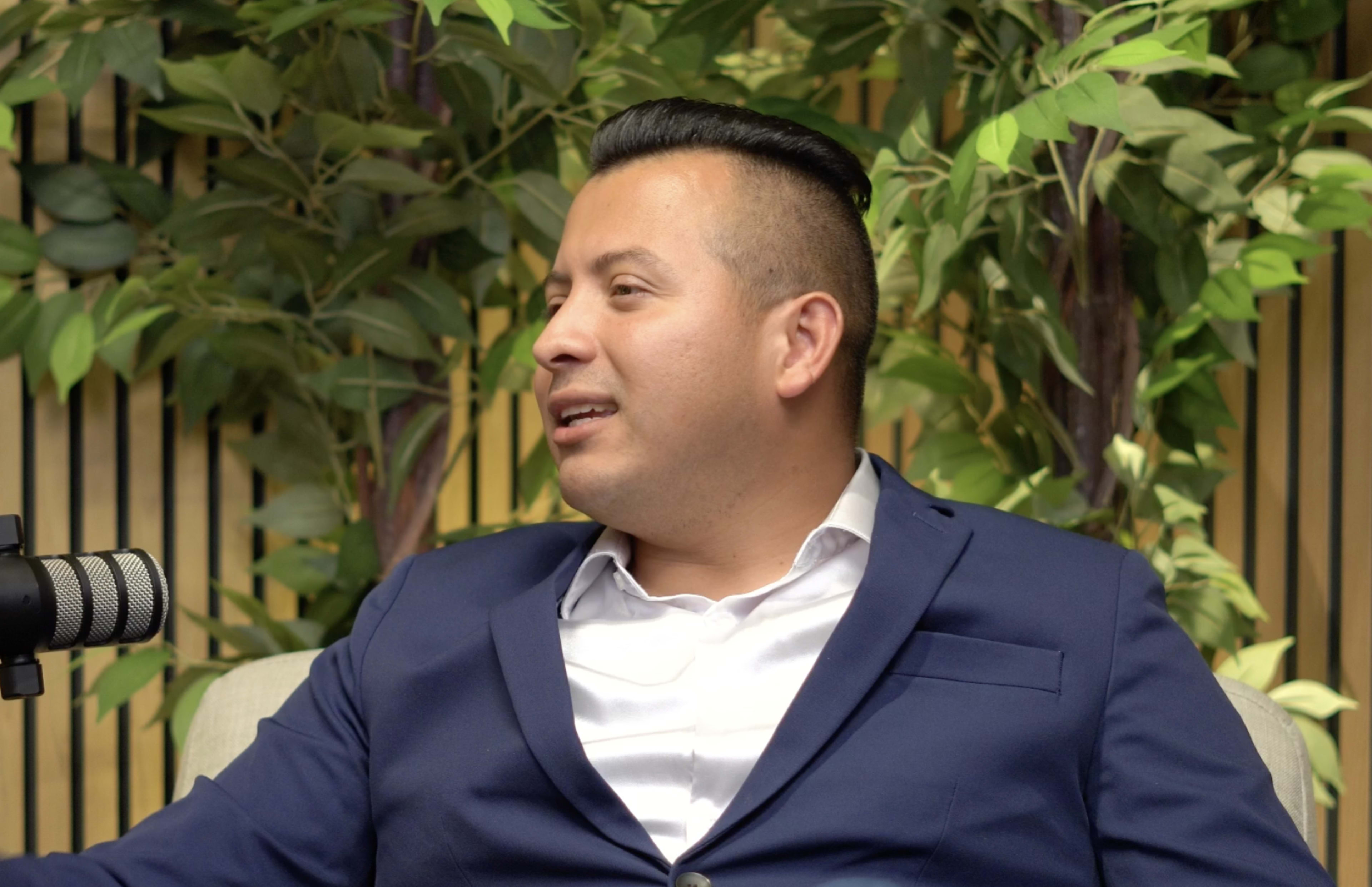 A man in a navy suit sits on a couch next to a microphone, with greenery in the background.