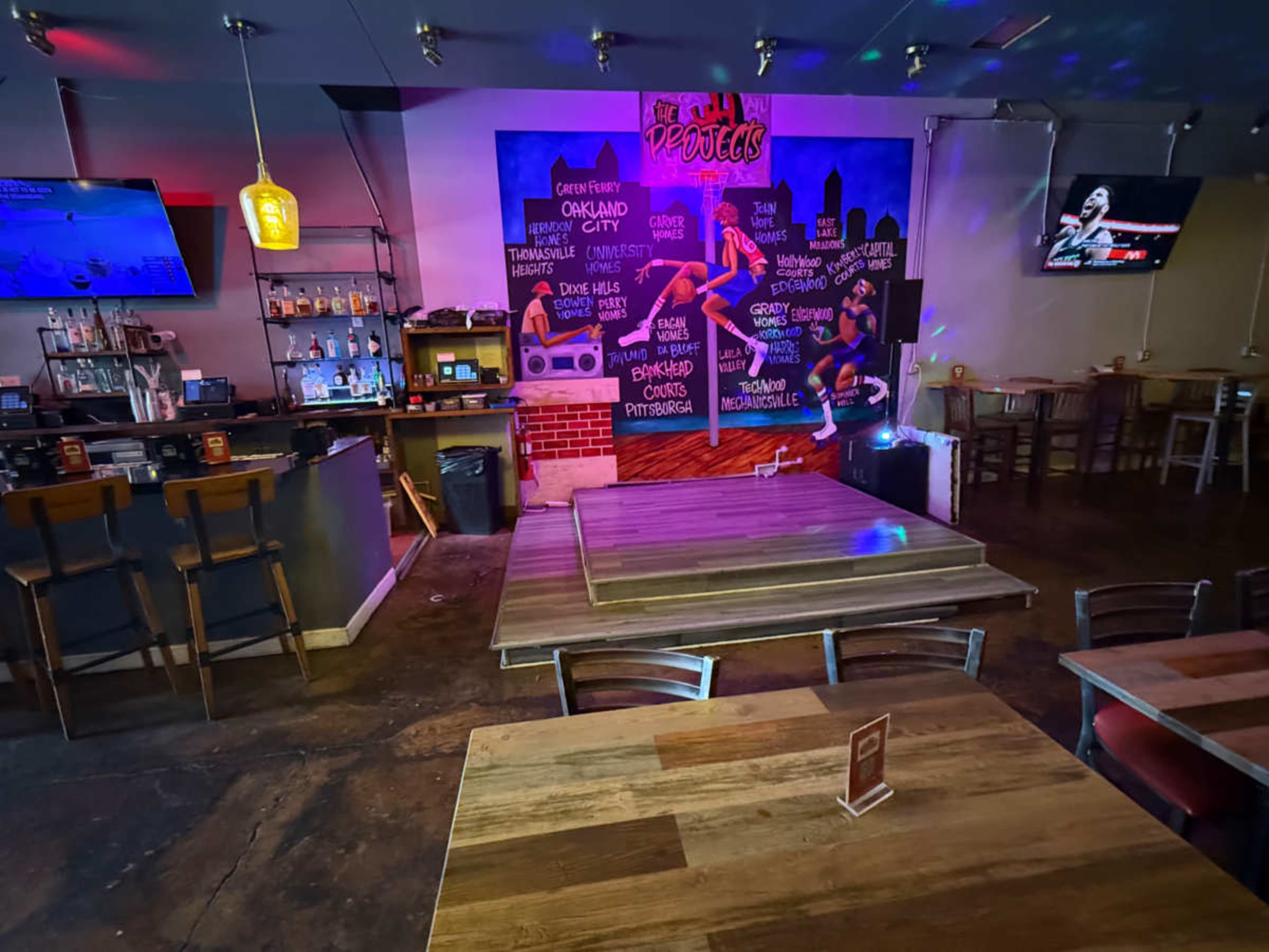 The image shows a bar area with a colorful mural on the wall, featuring various beverage and food options, and several wooden tables arranged nearby.