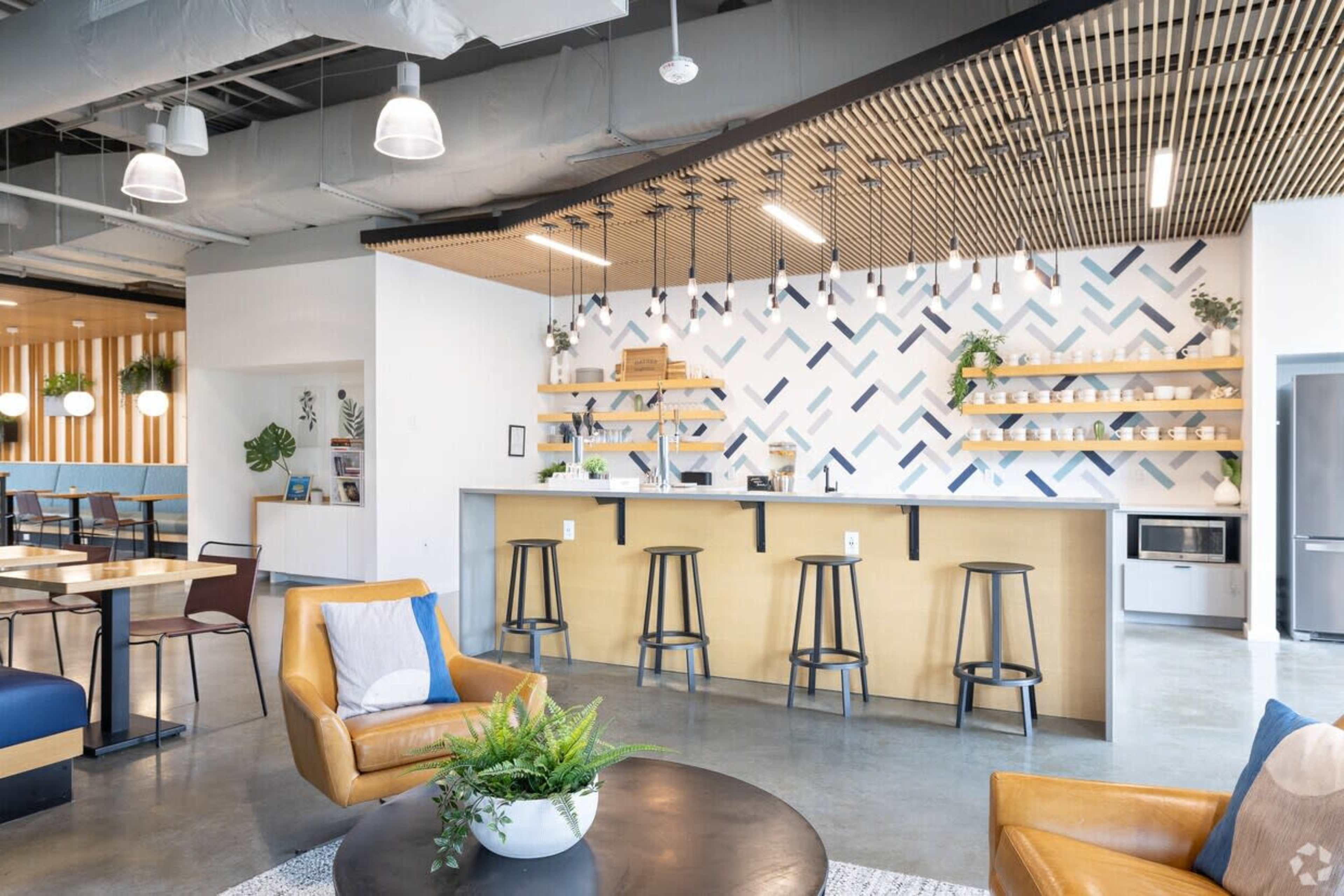 The image shows a modern, open-plan café area featuring a bar with stools, decorative wall patterns, indoor plants, and seating arrangements with tables and chairs.