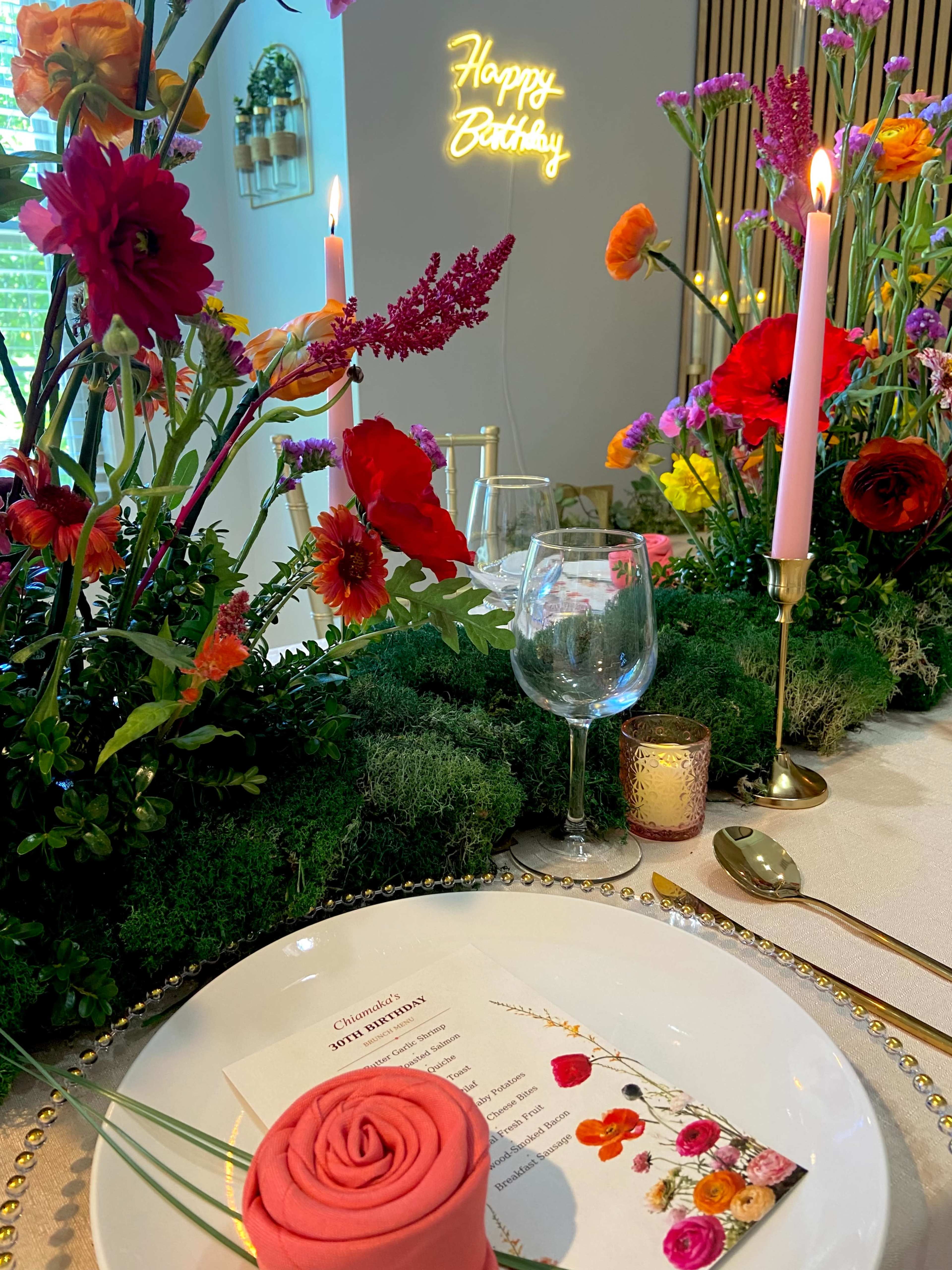 A beautifully set dining table is adorned with vibrant flowers, a pink birthday cake rose, a glass of wine, and a glowing "Happy Birthday" sign.