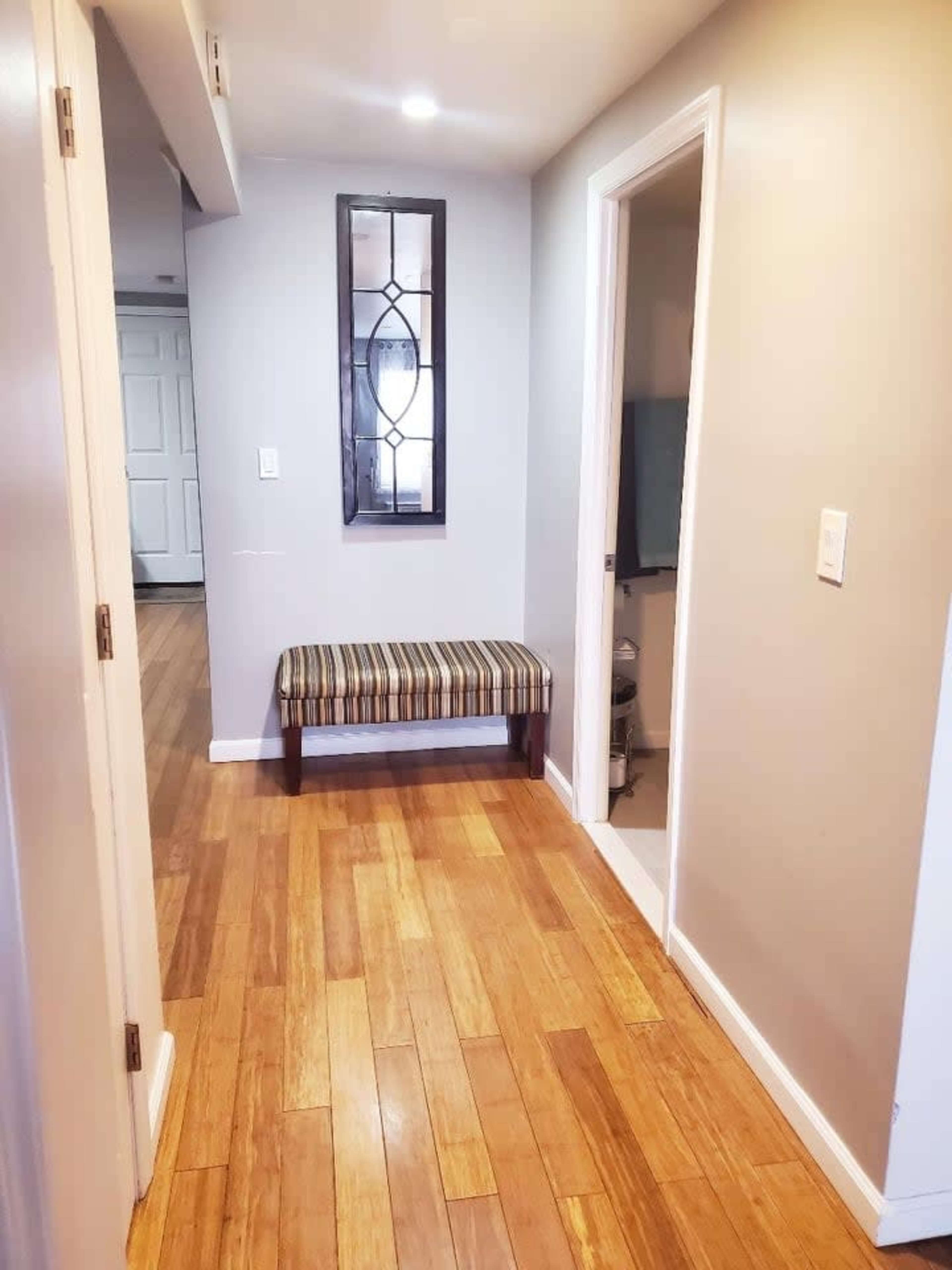 A narrow hallway with light hardwood flooring, featuring a striped bench against one wall and a mirror above it.
