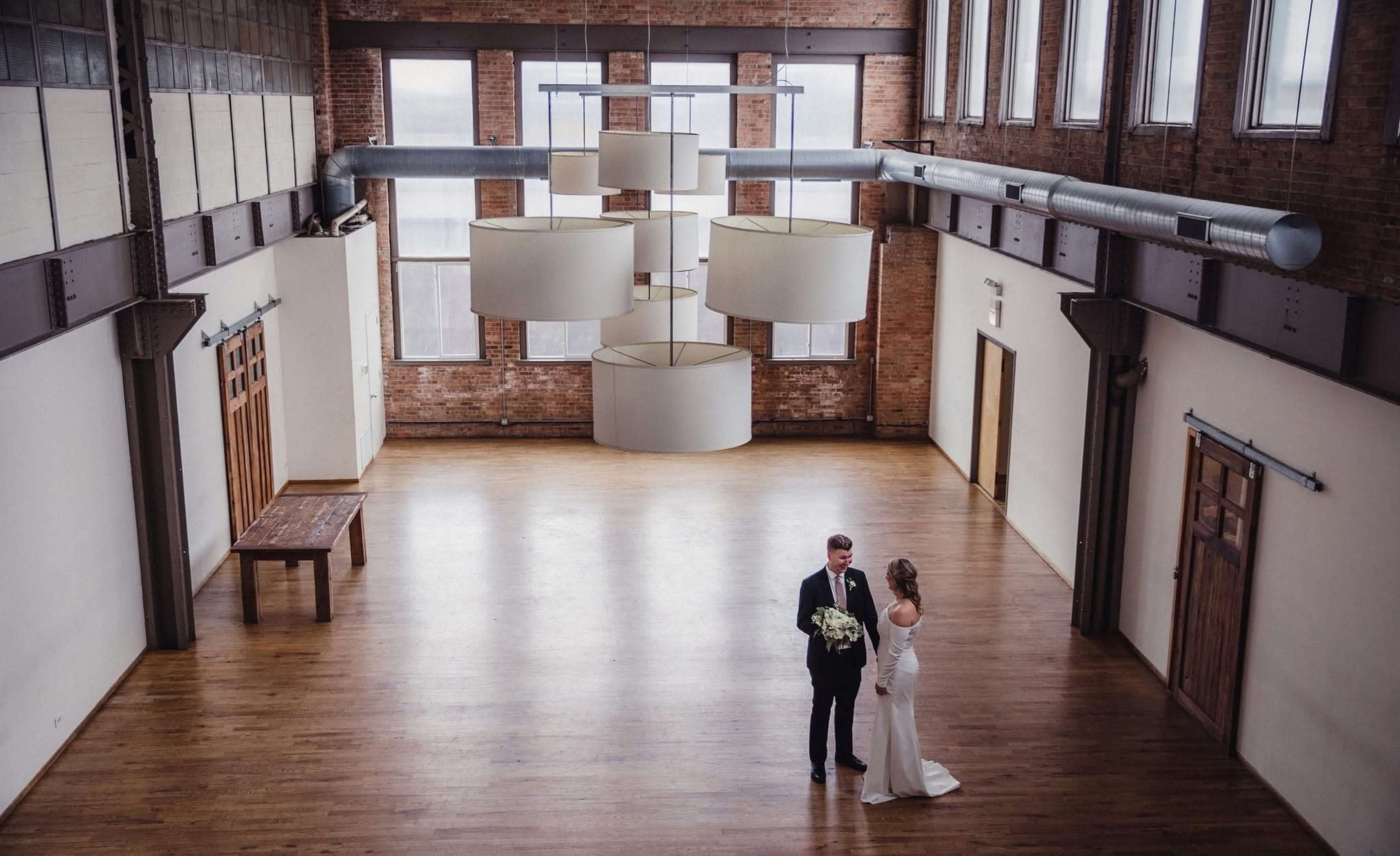 A couple stands facing each other in a spacious, softly lit room with wooden floors and large windows, featuring modern light fixtures hanging from the ceiling.