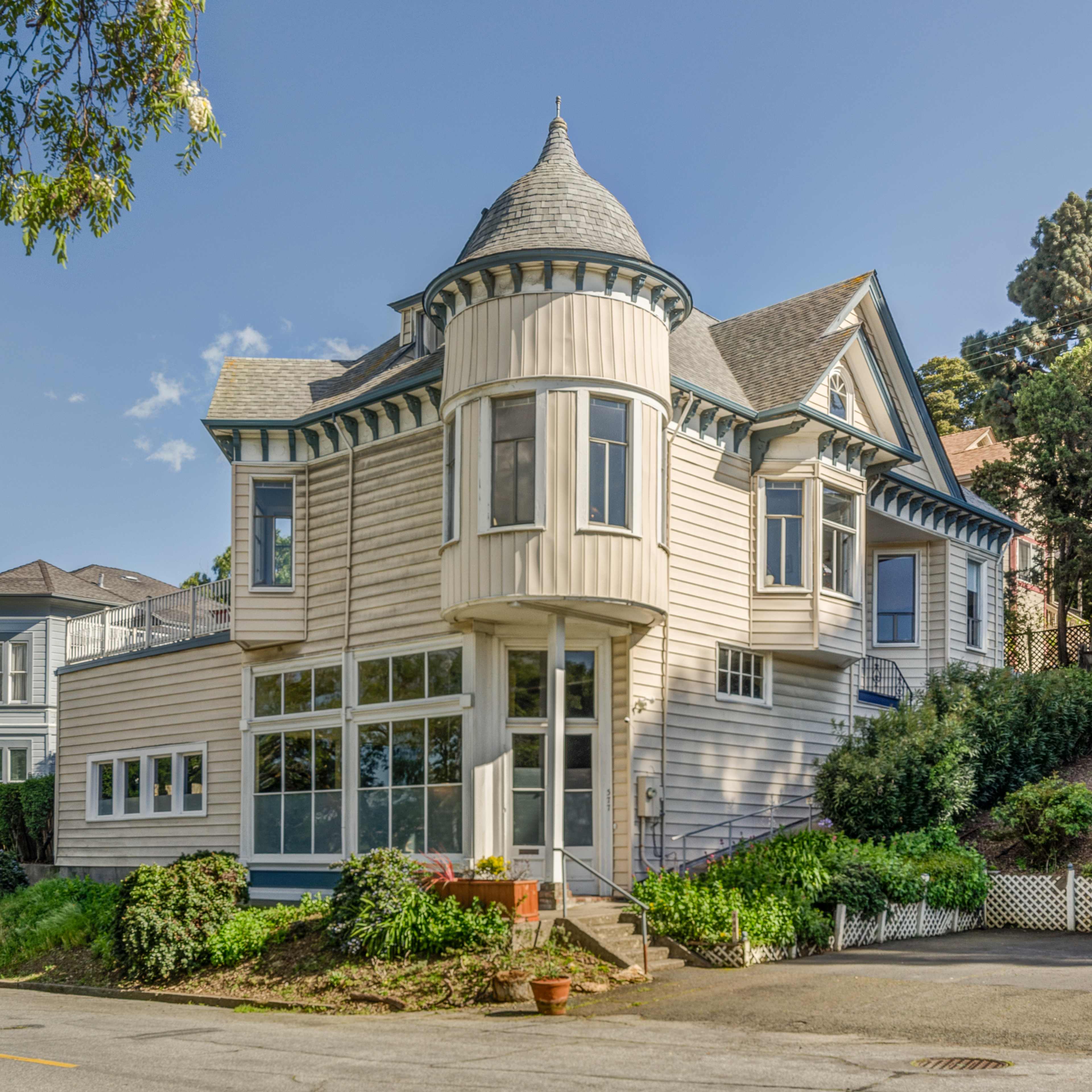 A two-story house with a conical turret and a large front porch, surrounded by greenery and situated at the corner of a street.