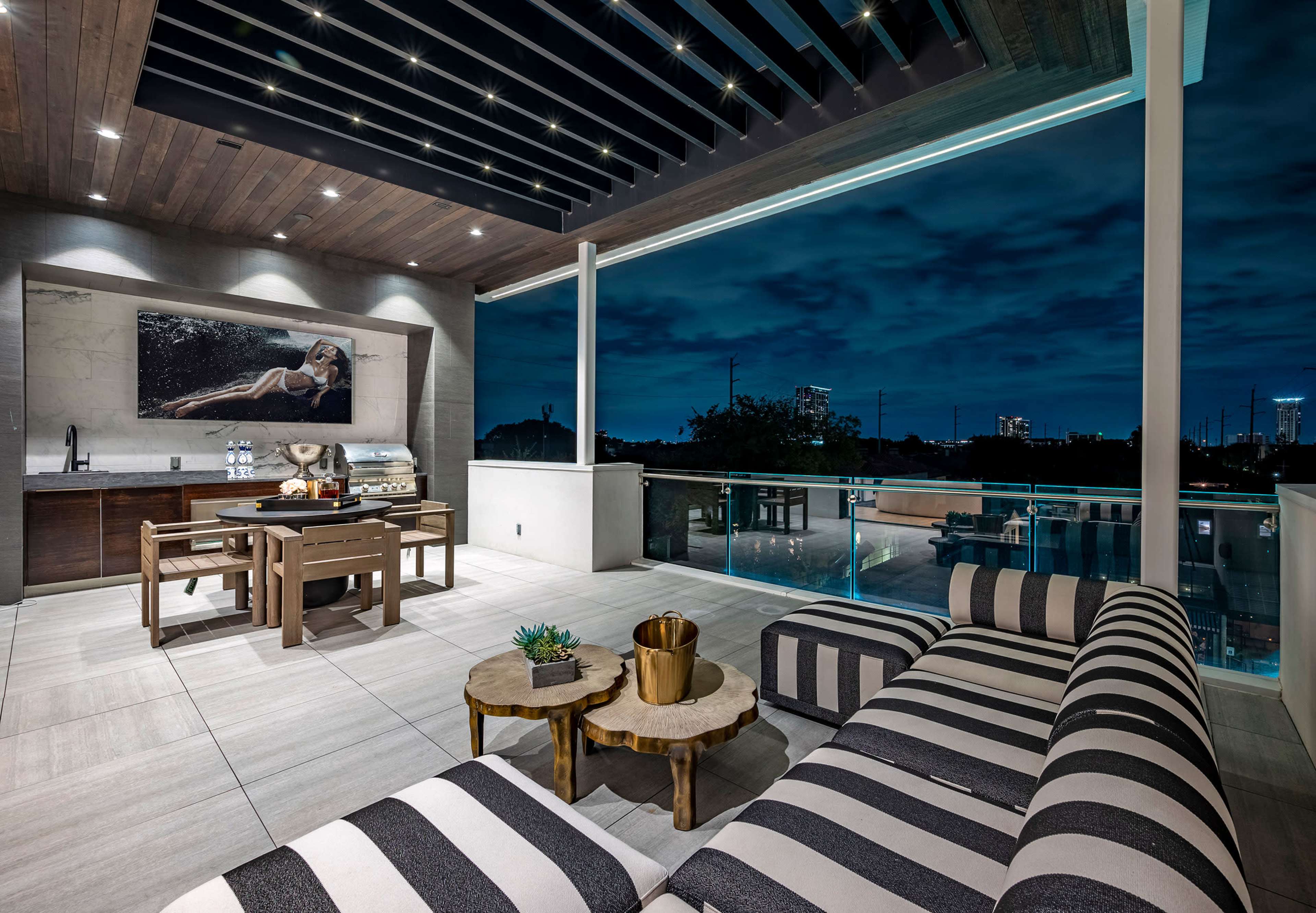 A modern outdoor patio with a striped sofa, dining area, and a view of a city skyline under a night sky.
