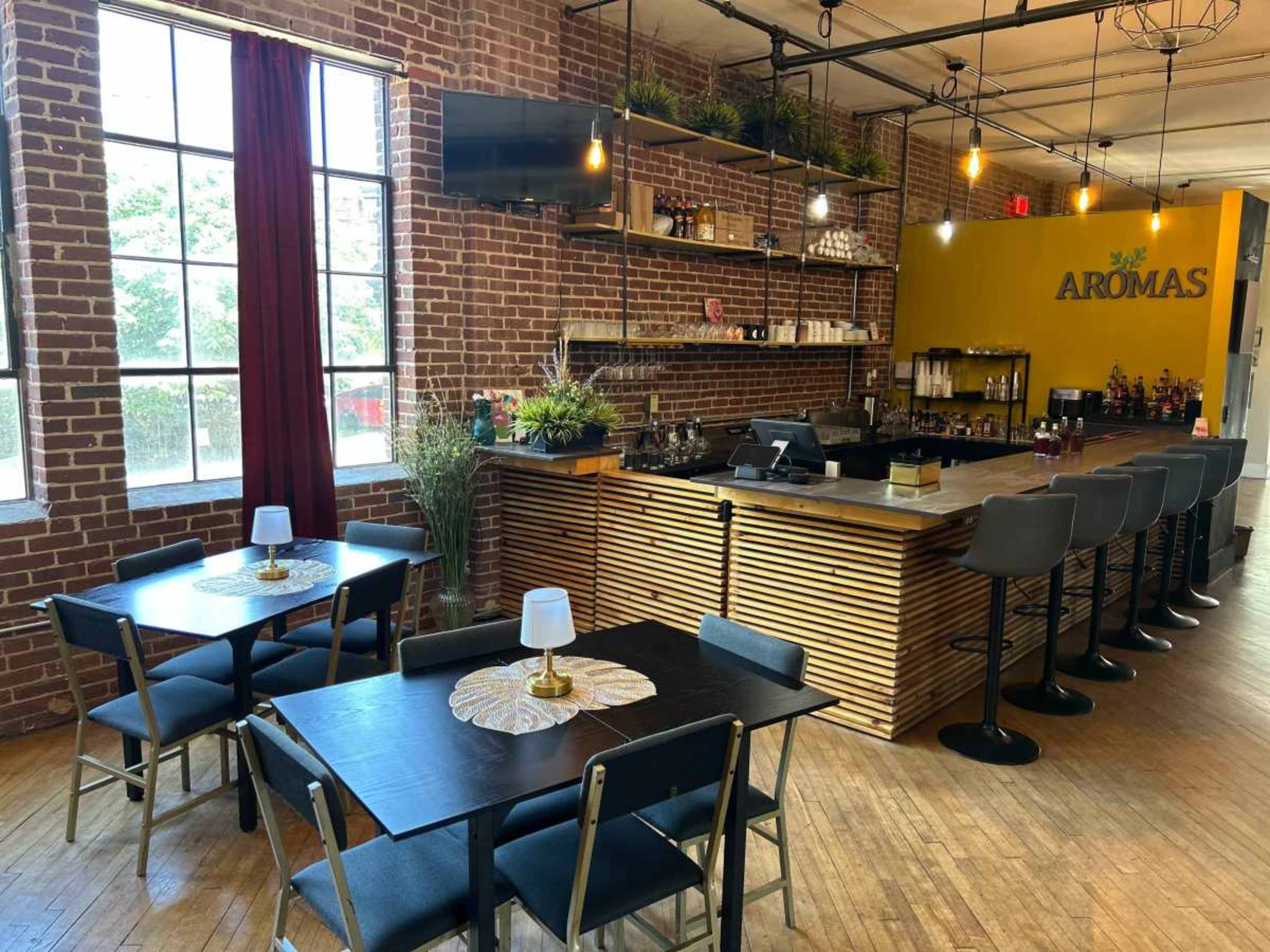 The image shows a modern café interior with brick walls, a wooden bar, and several tables with chairs arranged around the space.