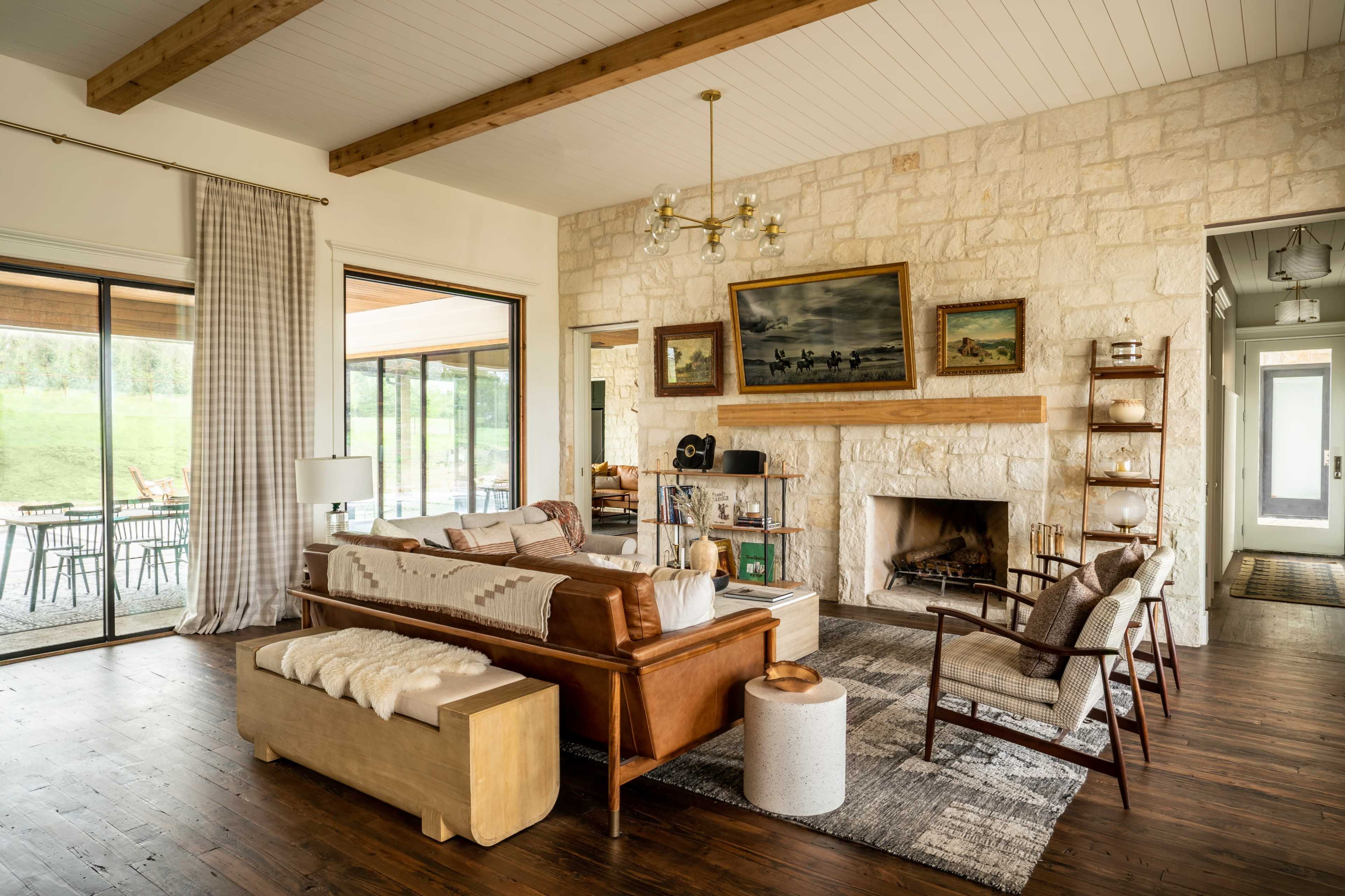 A modern living room features a stone fireplace, wooden beams, and a leather sofa, with large windows offering views of the outdoor patio.