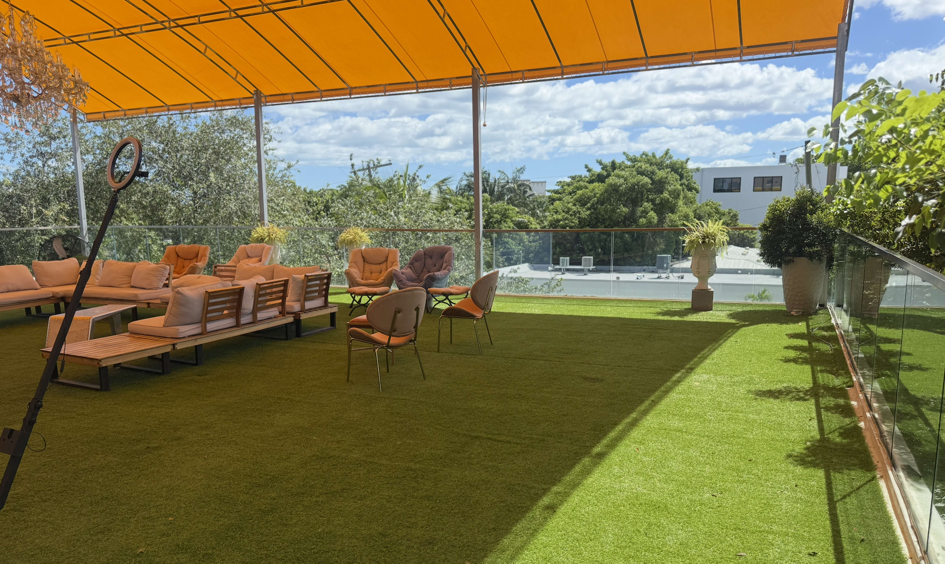 The image shows a rooftop terrace with orange awnings, cushioned seating, and potted plants against a backdrop of clouds and trees.
